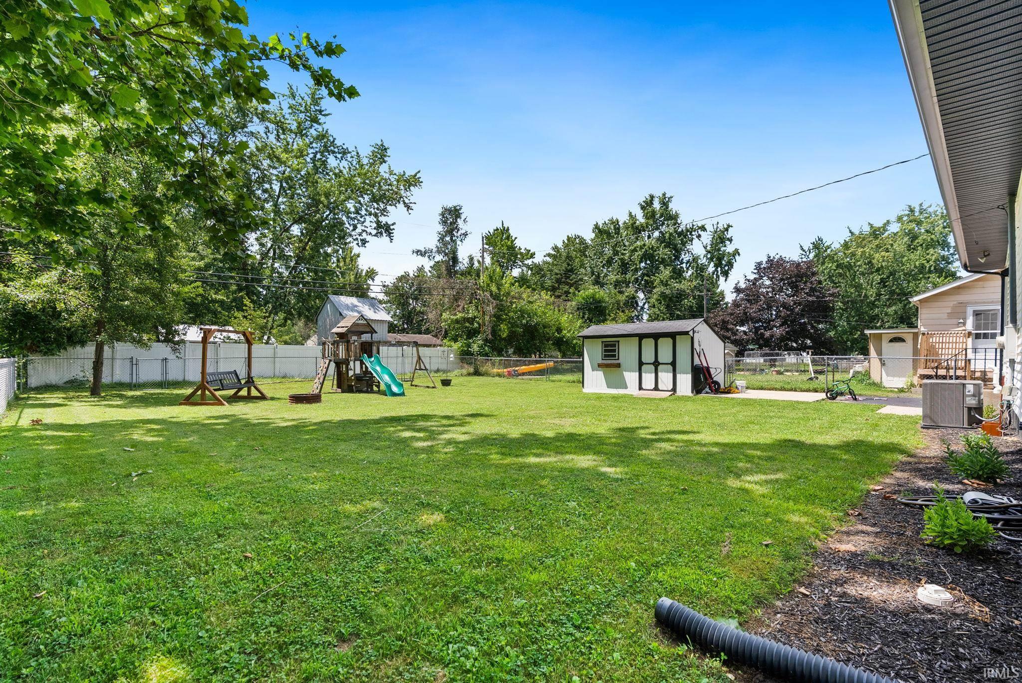 Expansive green lawn with mature trees, featuring a white privacy fence, a storage shed, and a concrete patio area