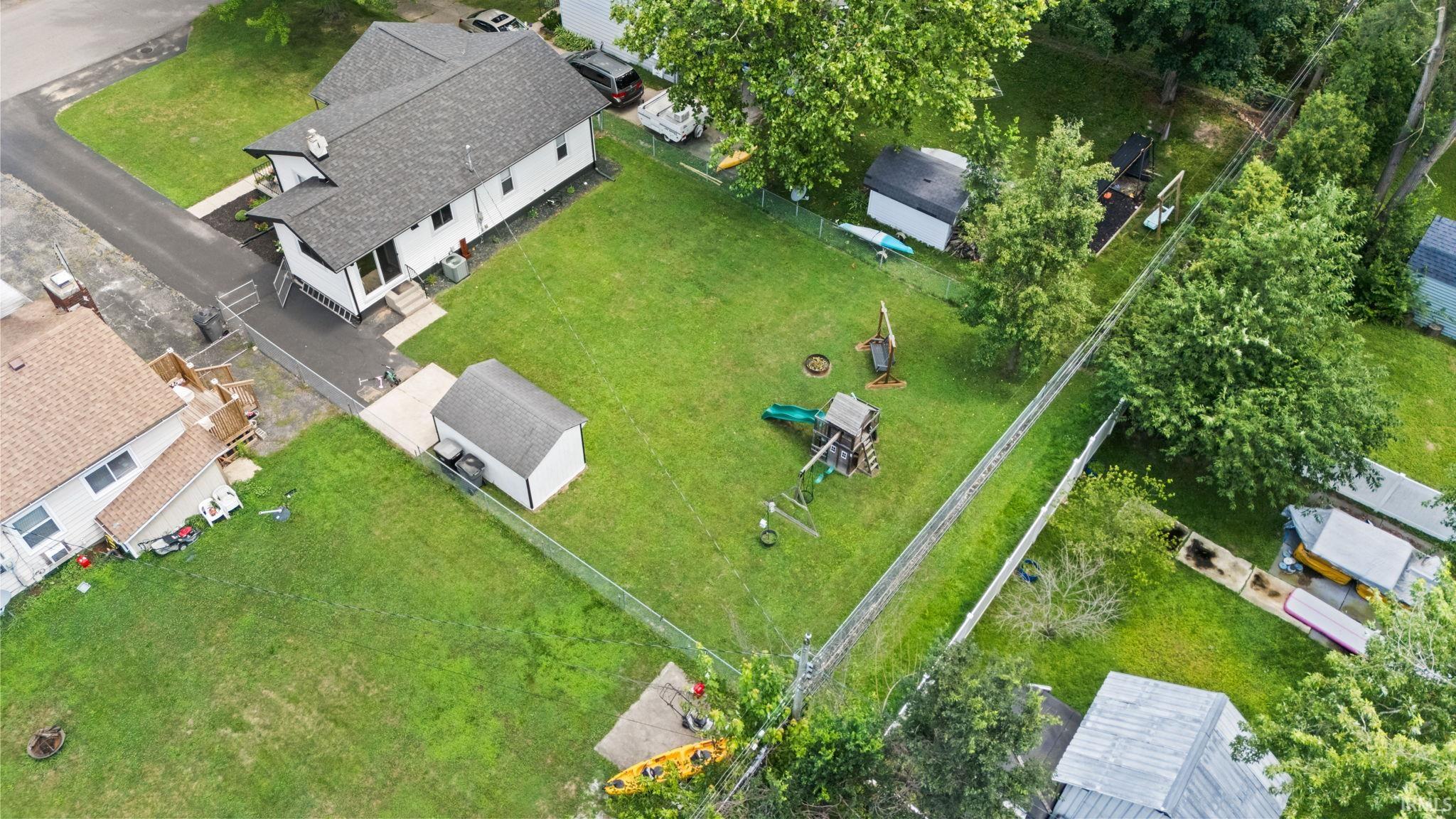Spacious property featuring a white exterior residence with a dark shingle roof, paved driveway, expansive lawn, and two detached storage structures