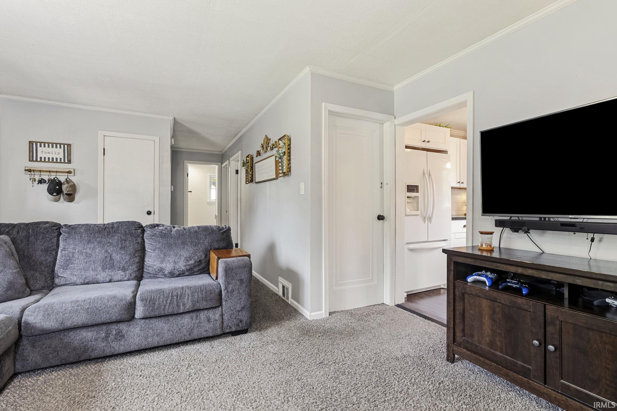 Carpeted living area featuring light gray walls, white trim, and a doorway opening to a kitchen with white appliances