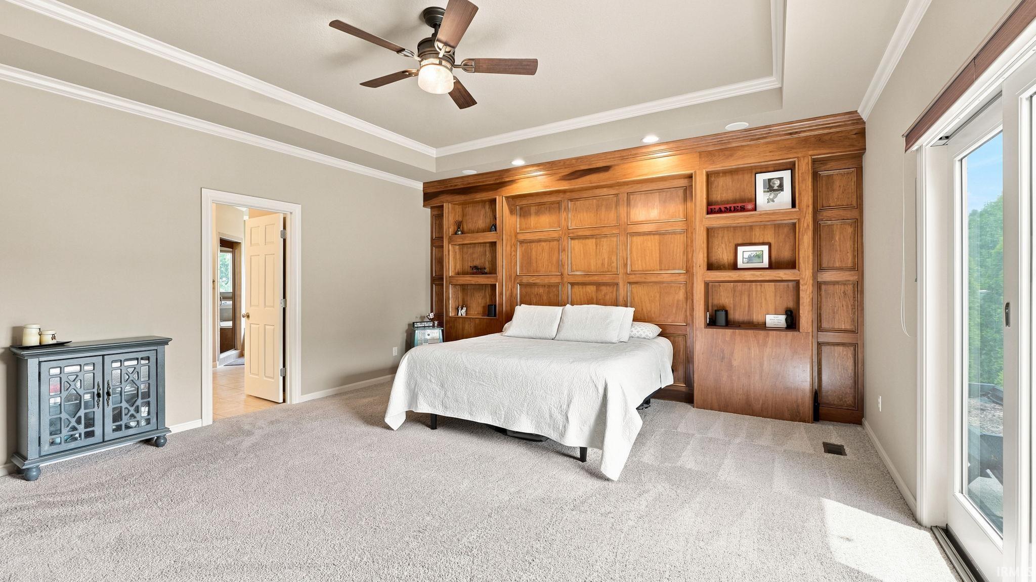 Spacious bedroom featuring light-toned carpeting, a built-in wood-finish wall unit with shelving, a ceiling fan with integrated lighting, a tray ceiling, and glass-paneled double doors providing outdoor access