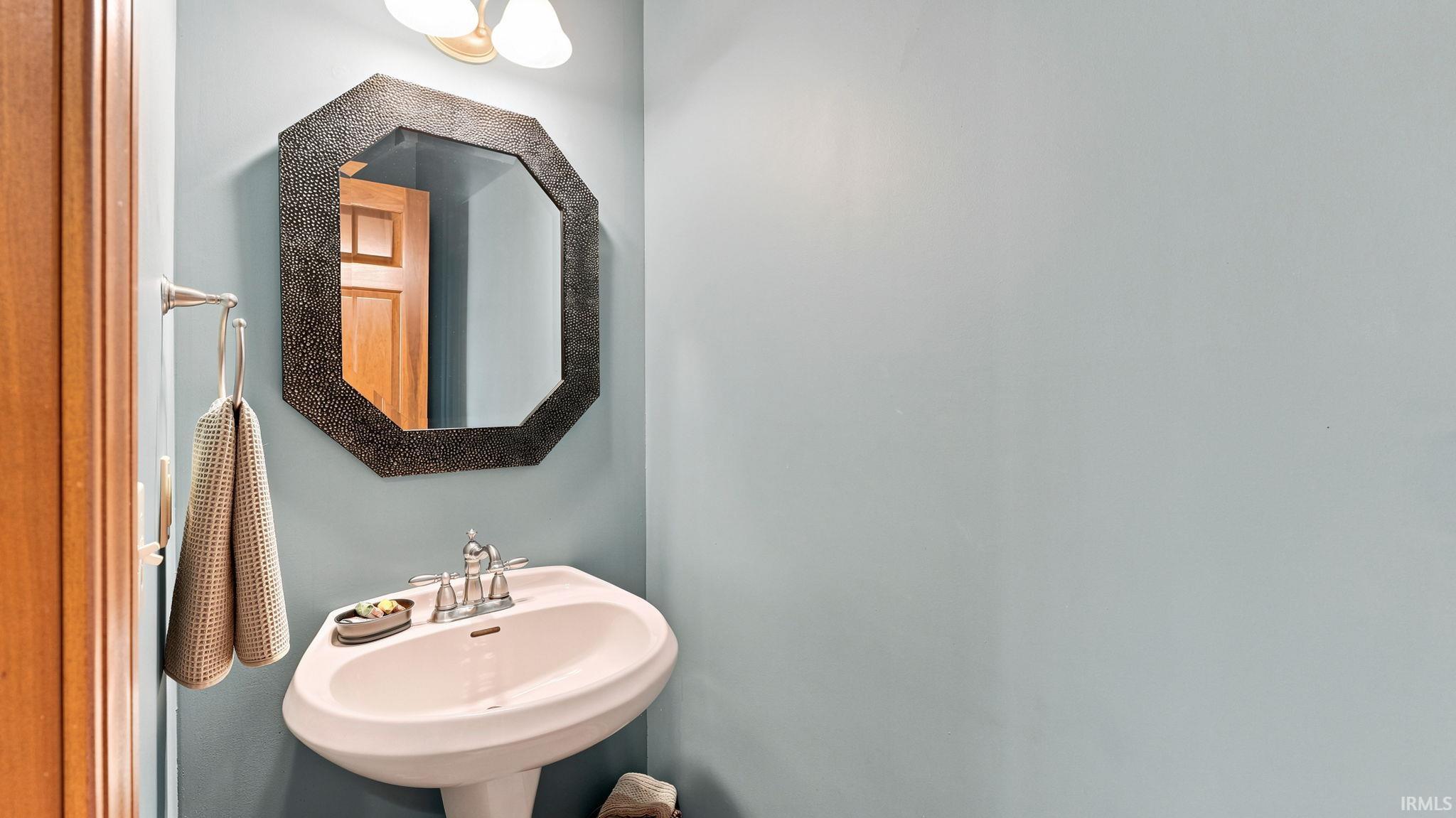 Pedestal sink with chrome faucet, complemented by an octagonal framed mirror and a wall-mounted light fixture