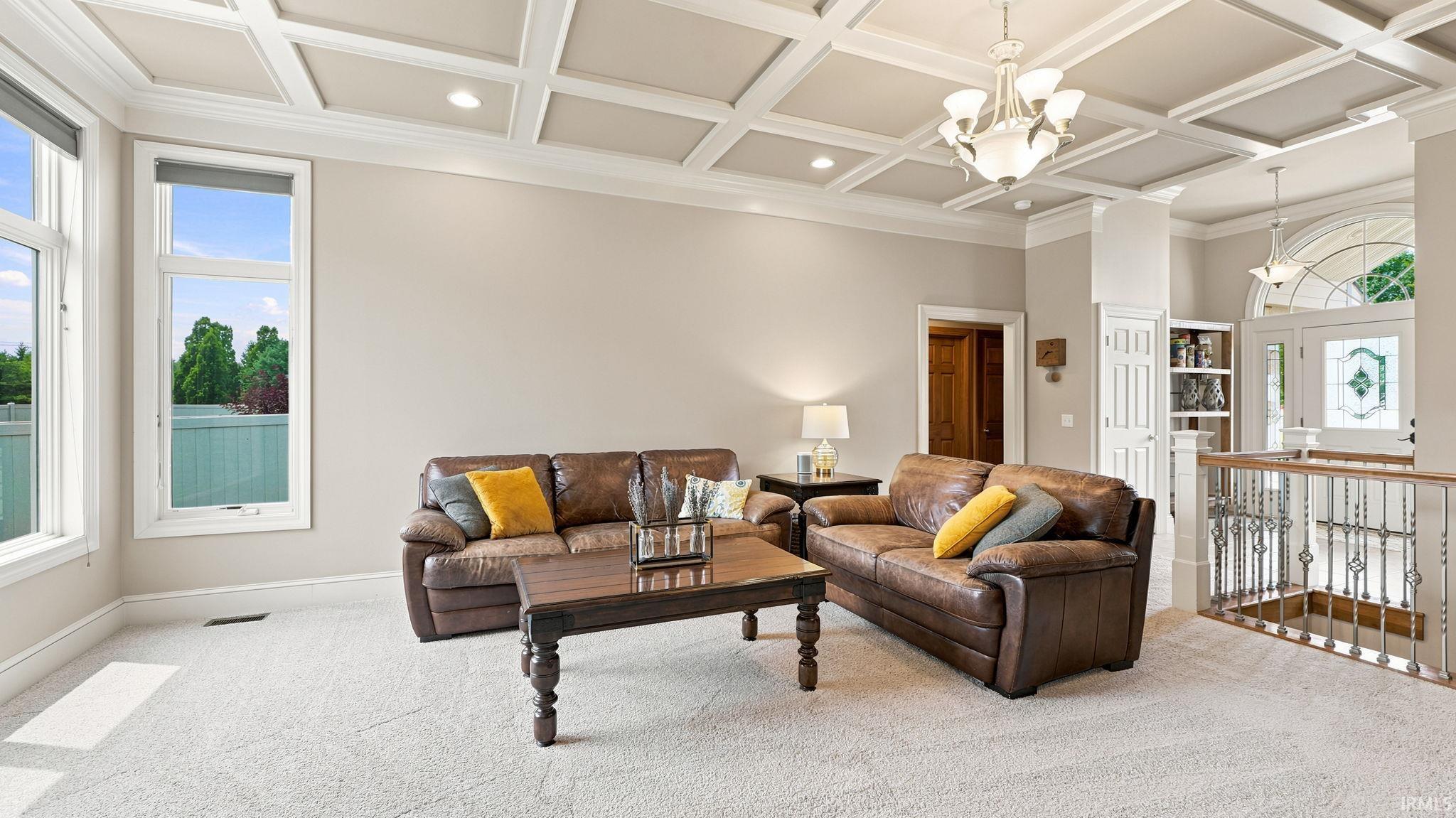 Spacious living area featuring a coffered ceiling with recessed lighting, large windows, light-colored carpeting, a decorative railing, and an elegant chandelier