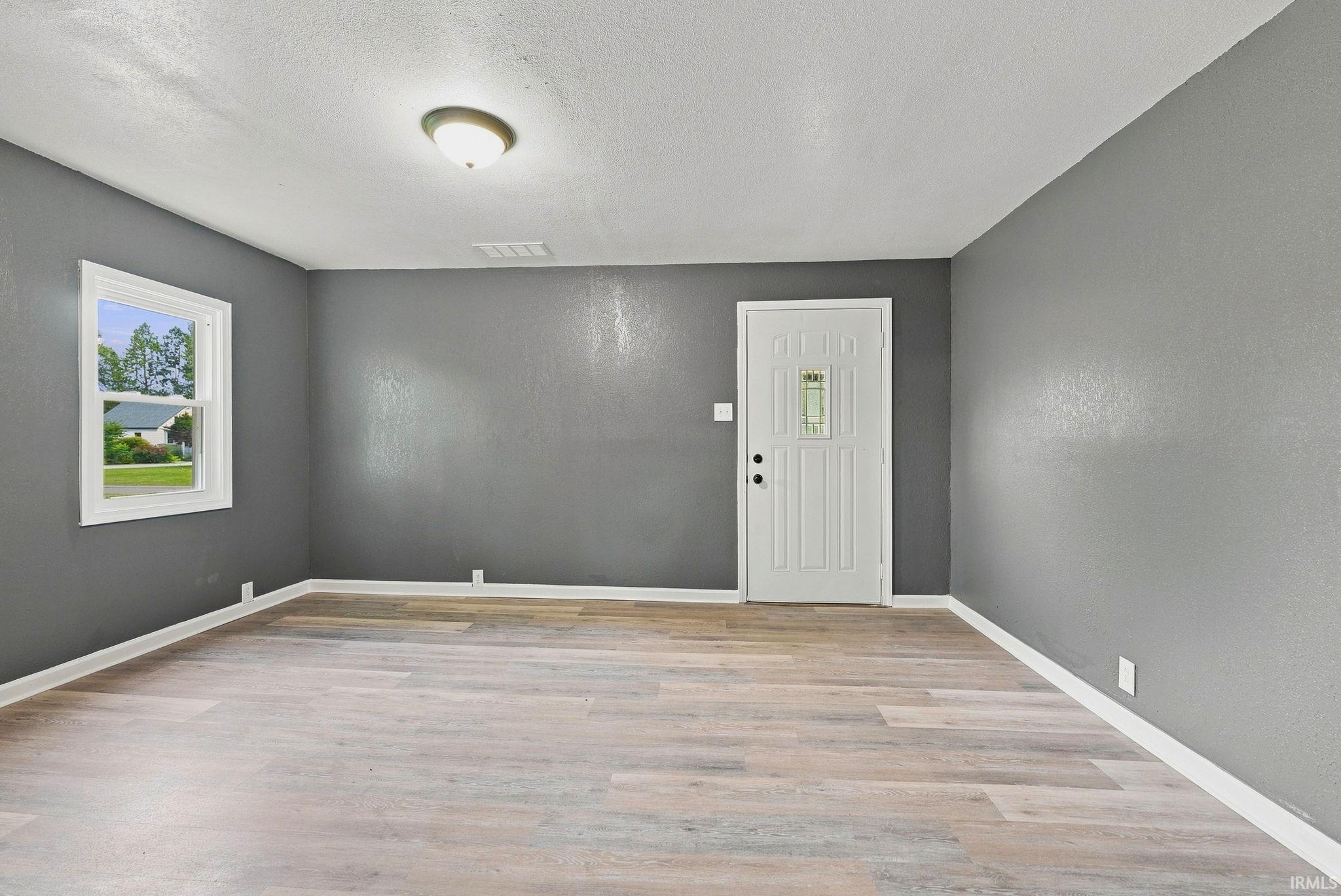Spacious room featuring wood-finish flooring, neutral gray walls, a white entry door with a decorative glass insert, a white-framed window, and a ceiling-mounted light fixture