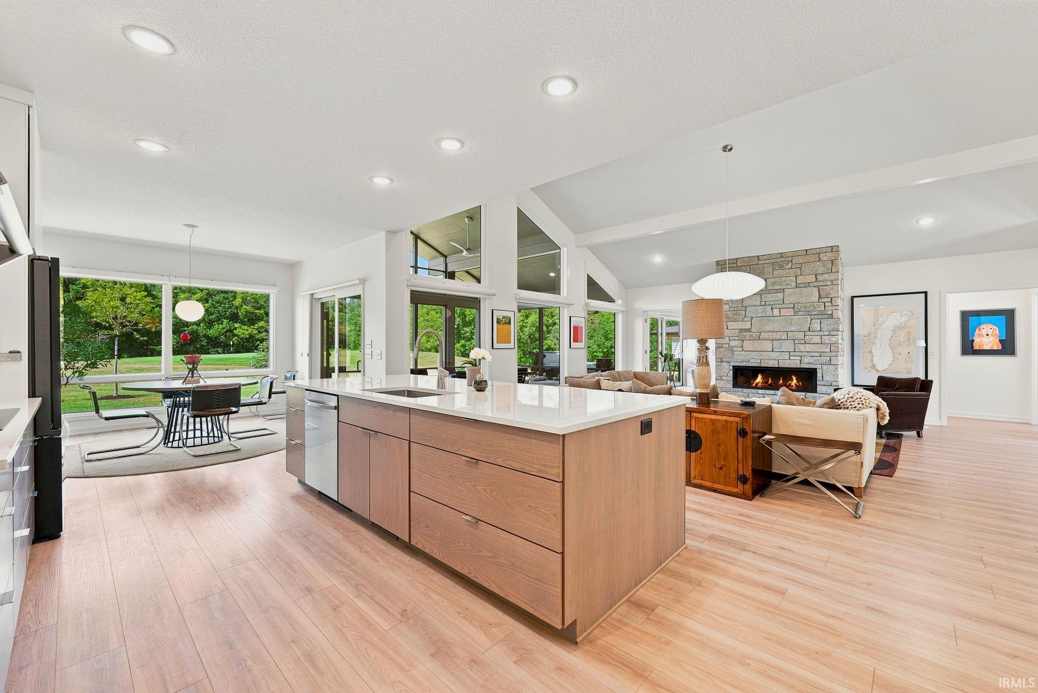 Bright open-concept interior featuring light wood-finish flooring, a large kitchen island with a light-toned countertop and integrated sink, stainless steel appliances, and a stone-faced fireplace