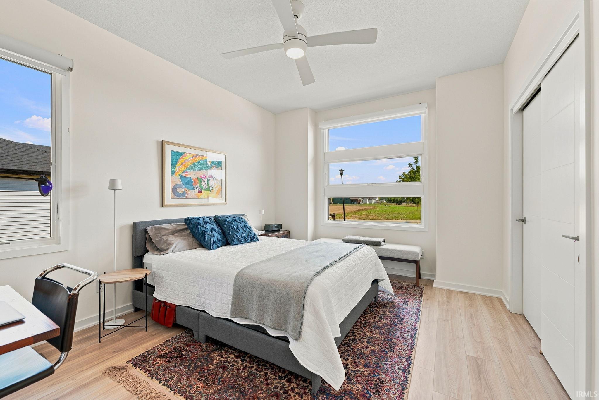 Bright room featuring light-toned wood-finish flooring, a contemporary ceiling fan, dual large windows, and a built-in closet with modern doors
