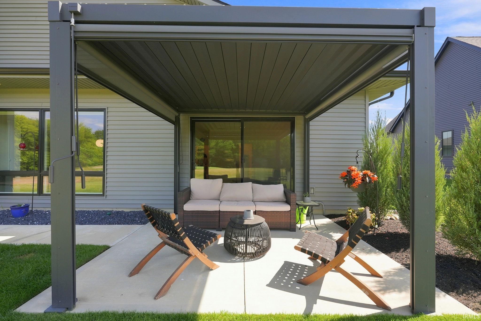 Expansive covered patio featuring a dark-toned pergola, concrete flooring, and surrounding green lawn