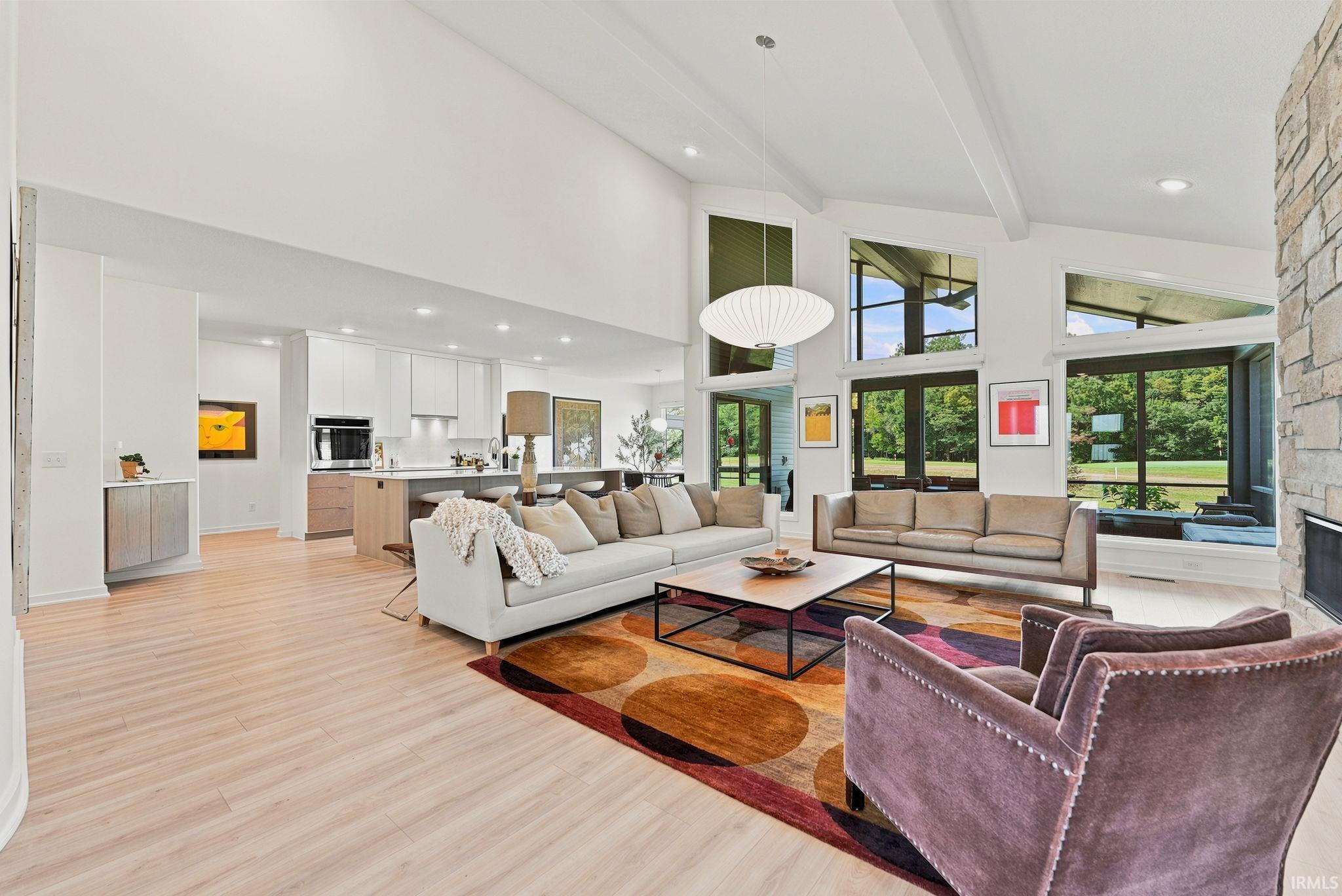 Spacious living area featuring high vaulted ceilings with exposed beams, wood-finish flooring, a stone-clad fireplace, and expansive windows