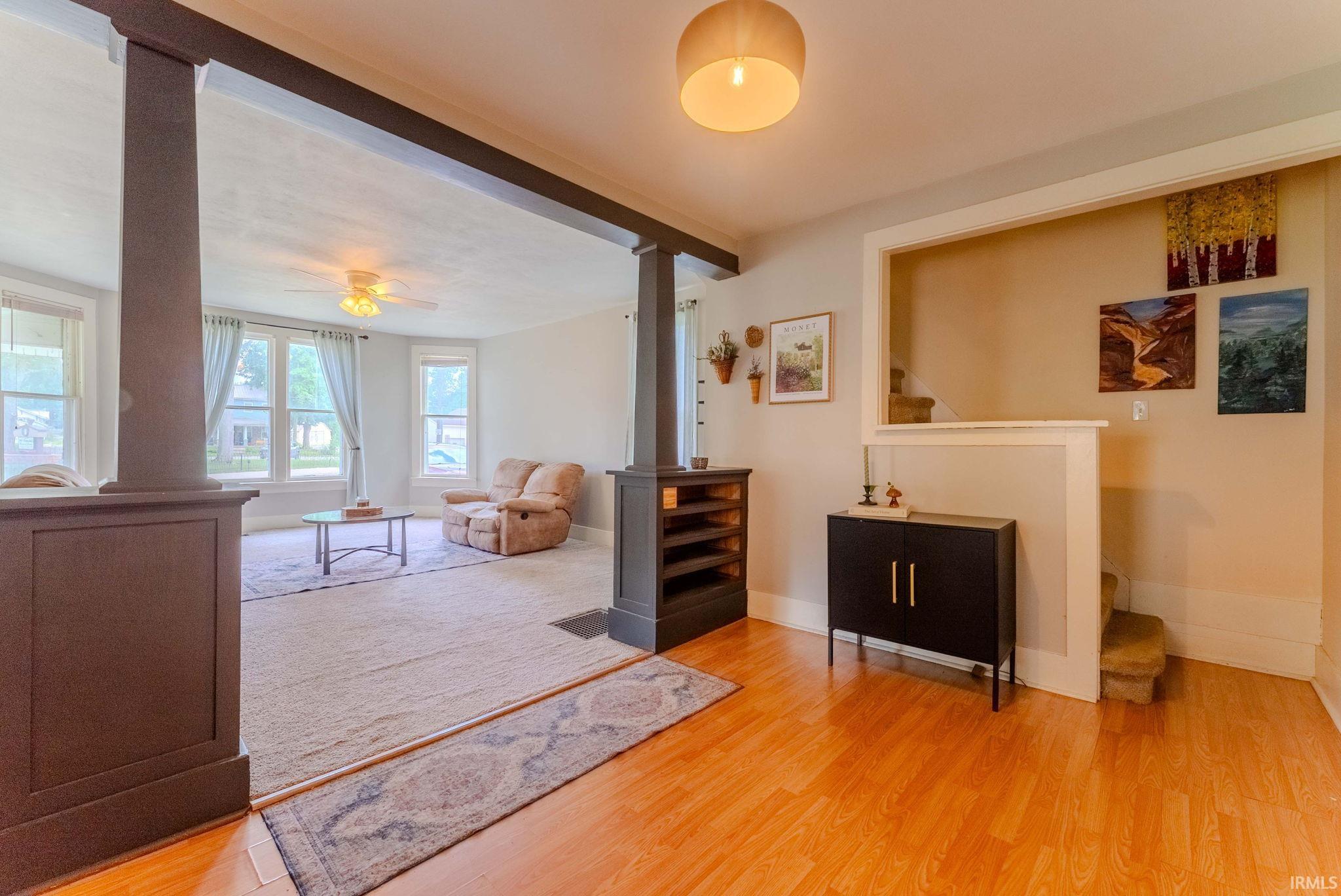 Open-concept living space featuring wood-finish flooring, a carpeted area, multiple large windows, a ceiling fan, and decorative structural columns