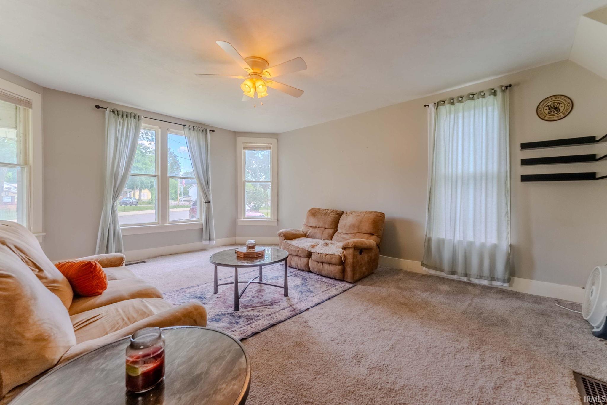 Spacious living area featuring light-colored wall paint, carpet flooring, and a ceiling fan with integrated lighting