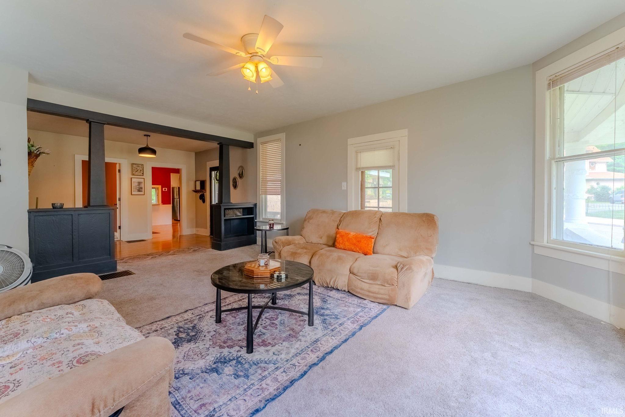 Spacious living area featuring light gray wall paint, light-colored carpeting, a ceiling fan with integrated lighting, and multiple windows