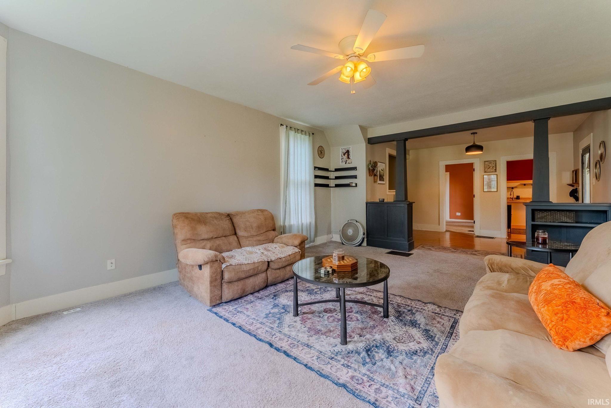 Carpeted living area featuring a ceiling fan with integrated lighting, a window with sheer coverings, and an open entryway with dark architectural columns and built-in shelving