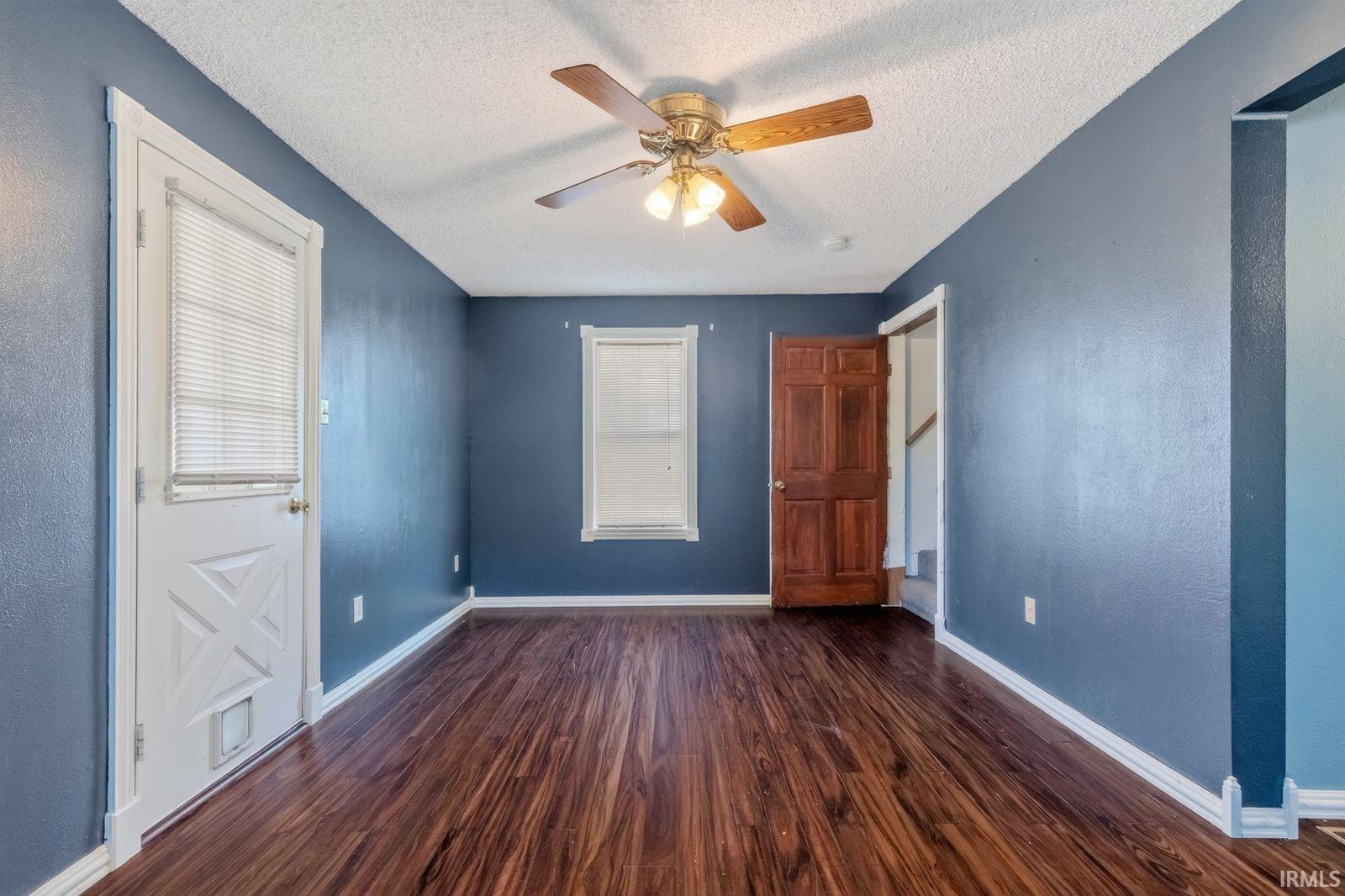 Spacious room featuring rich wood-finish flooring, a ceiling fan with integrated lighting, and a white door with a window blind
