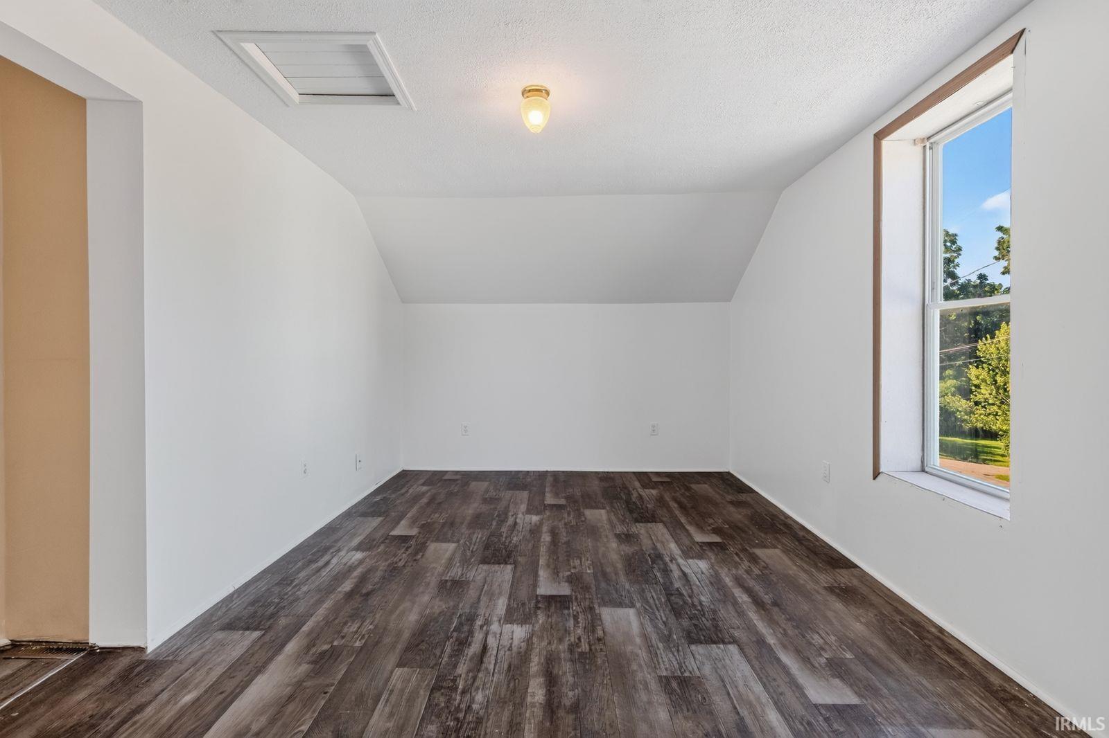 Spacious room featuring wood-finish flooring, crisp white walls, a single-pane window, and an attic access panel