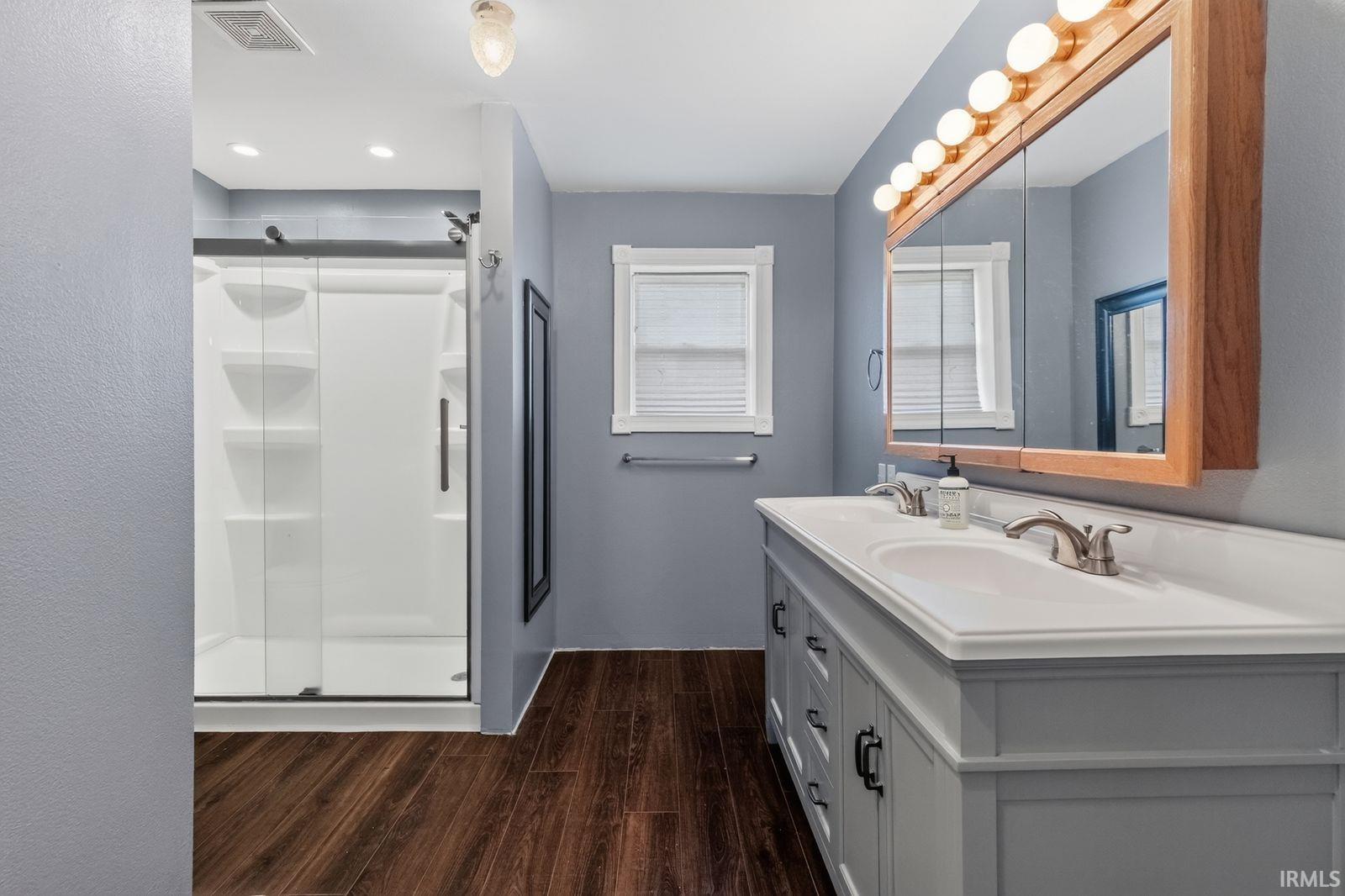 Bathroom featuring a dual-sink vanity with light-colored cabinetry and countertop, wood-finish flooring, and a glass-enclosed shower with a sliding door