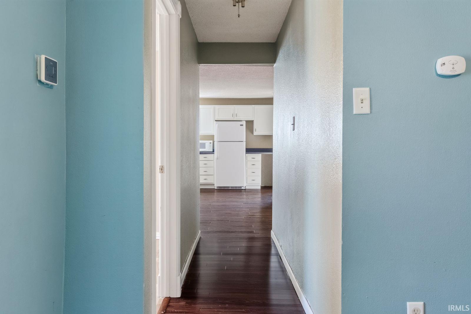 Hallway with wood-finish flooring extending to a kitchen featuring white cabinetry and appliances