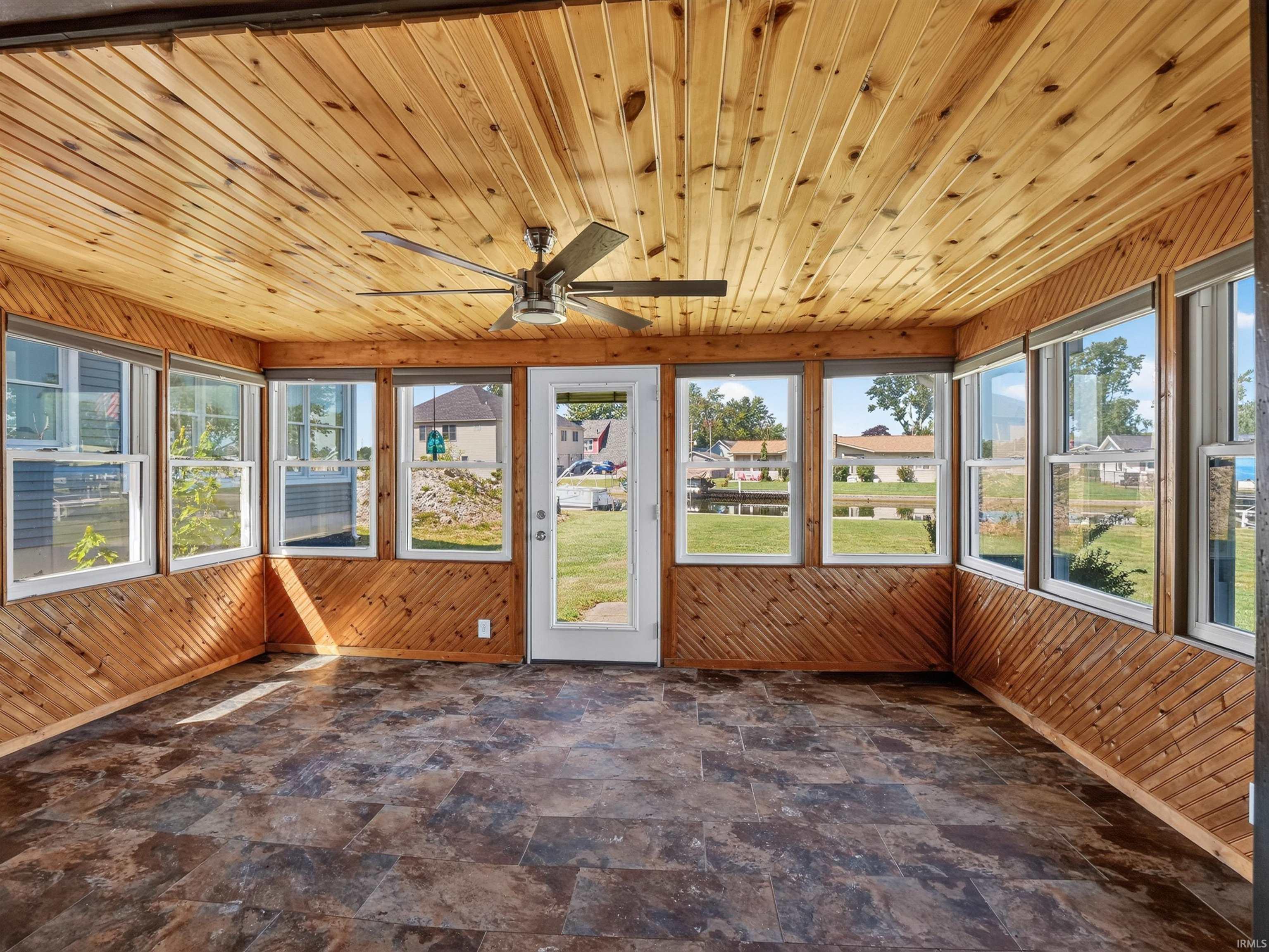 Sunroom featuring extensive natural wood plank paneling on walls and ceiling, a ceiling fan with light fixture, multiple windows, a glass-paneled exterior door, and patterned tile flooring