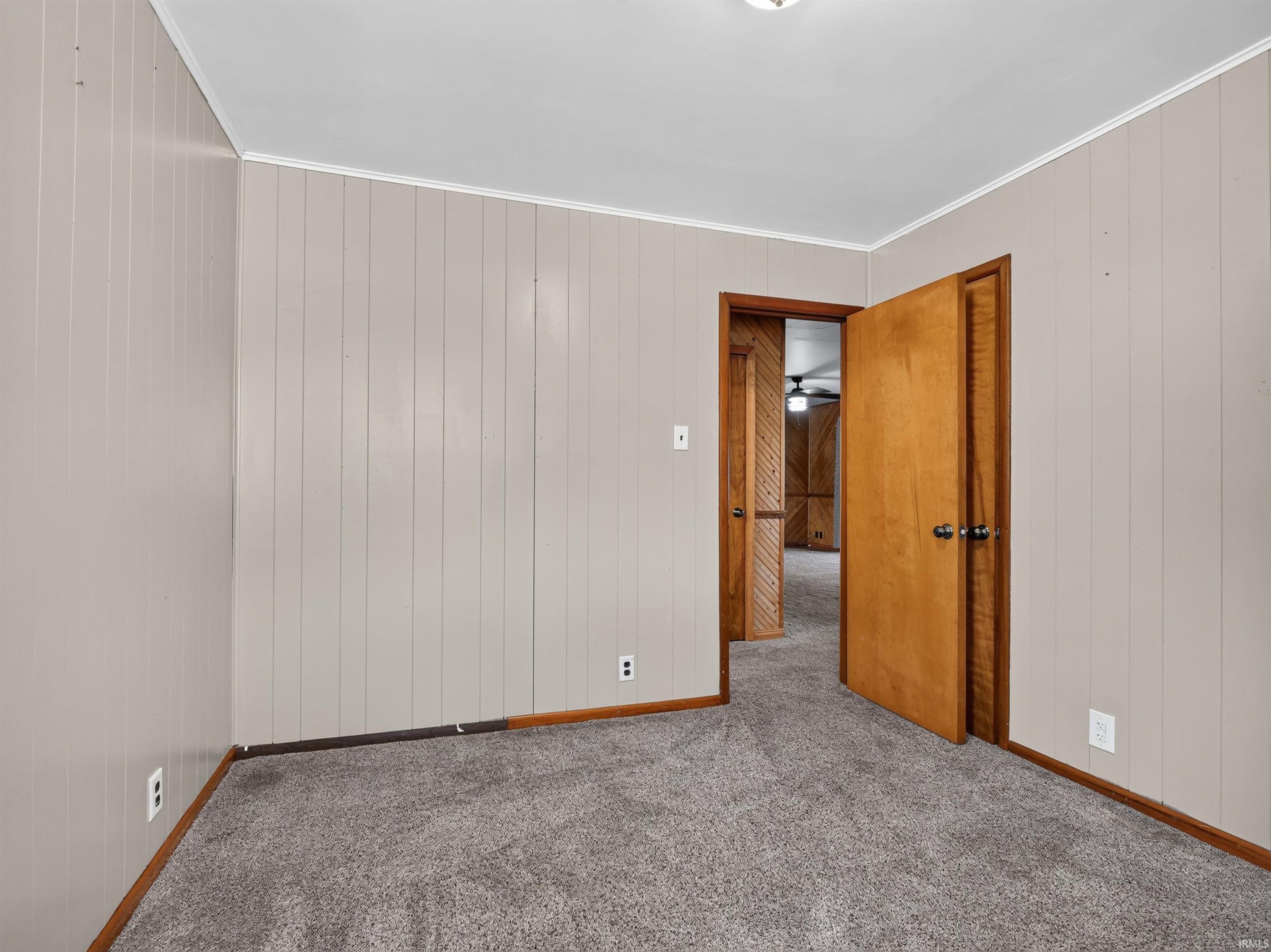 Room featuring vertical paneling walls, gray carpeting, natural wood trim, and a natural wood door with dark hardware