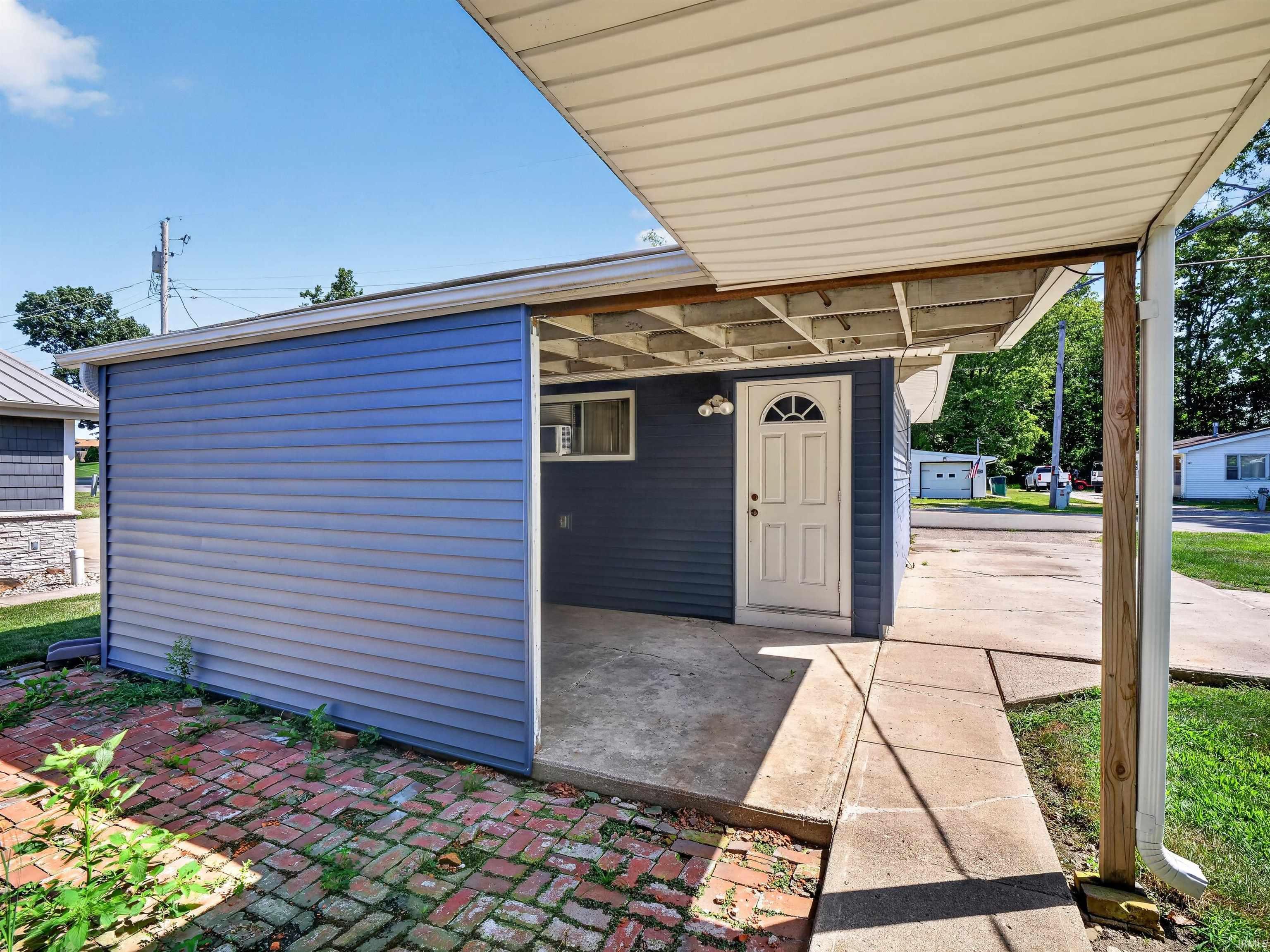 Covered patio area featuring a white entry door with an arched window, blue siding, and a brick patio surface