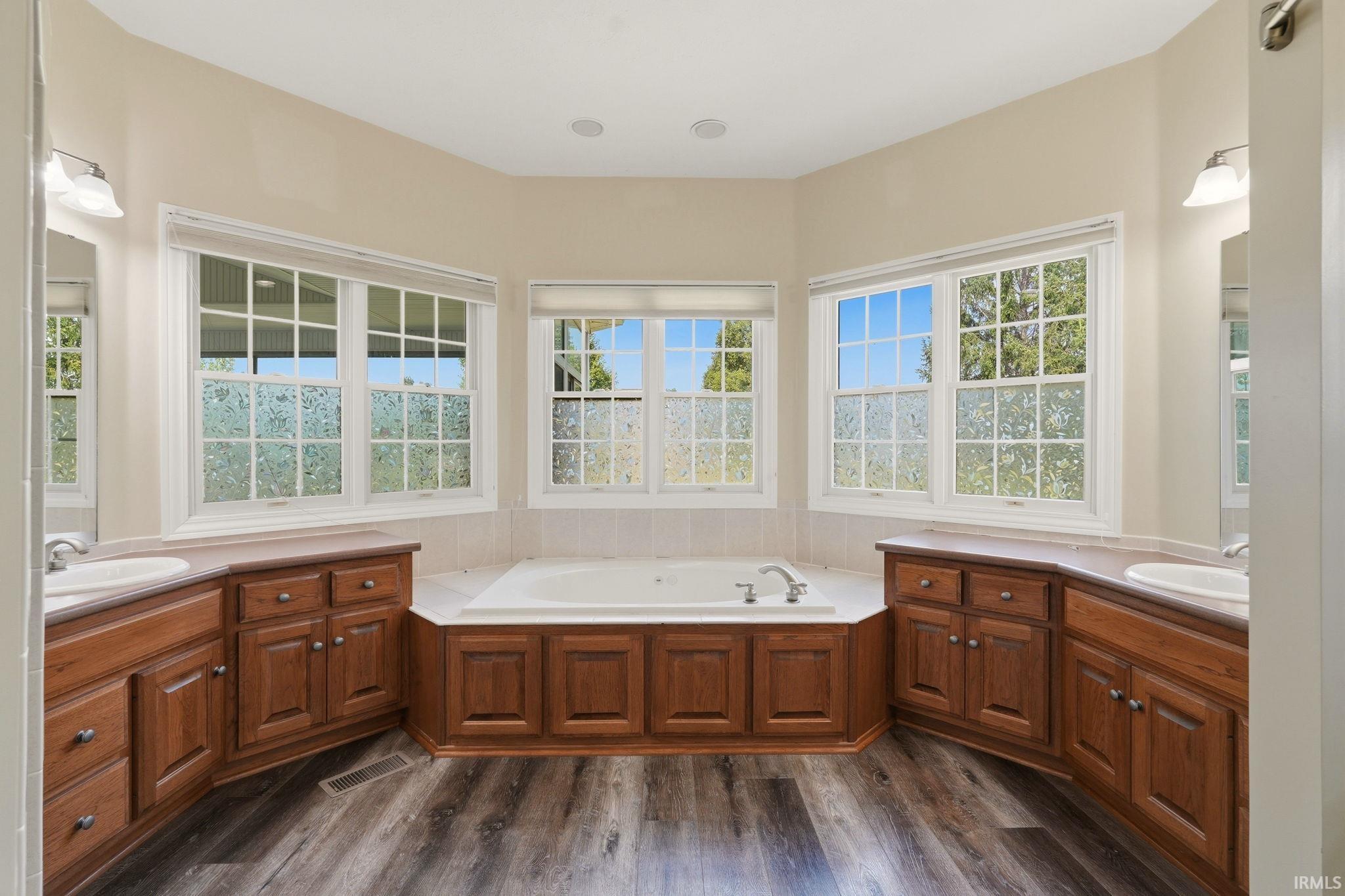 Spacious bathroom featuring a built-in soaking tub, dual vanity sinks, wood-finish cabinetry, and multiple windows with white grid patterns