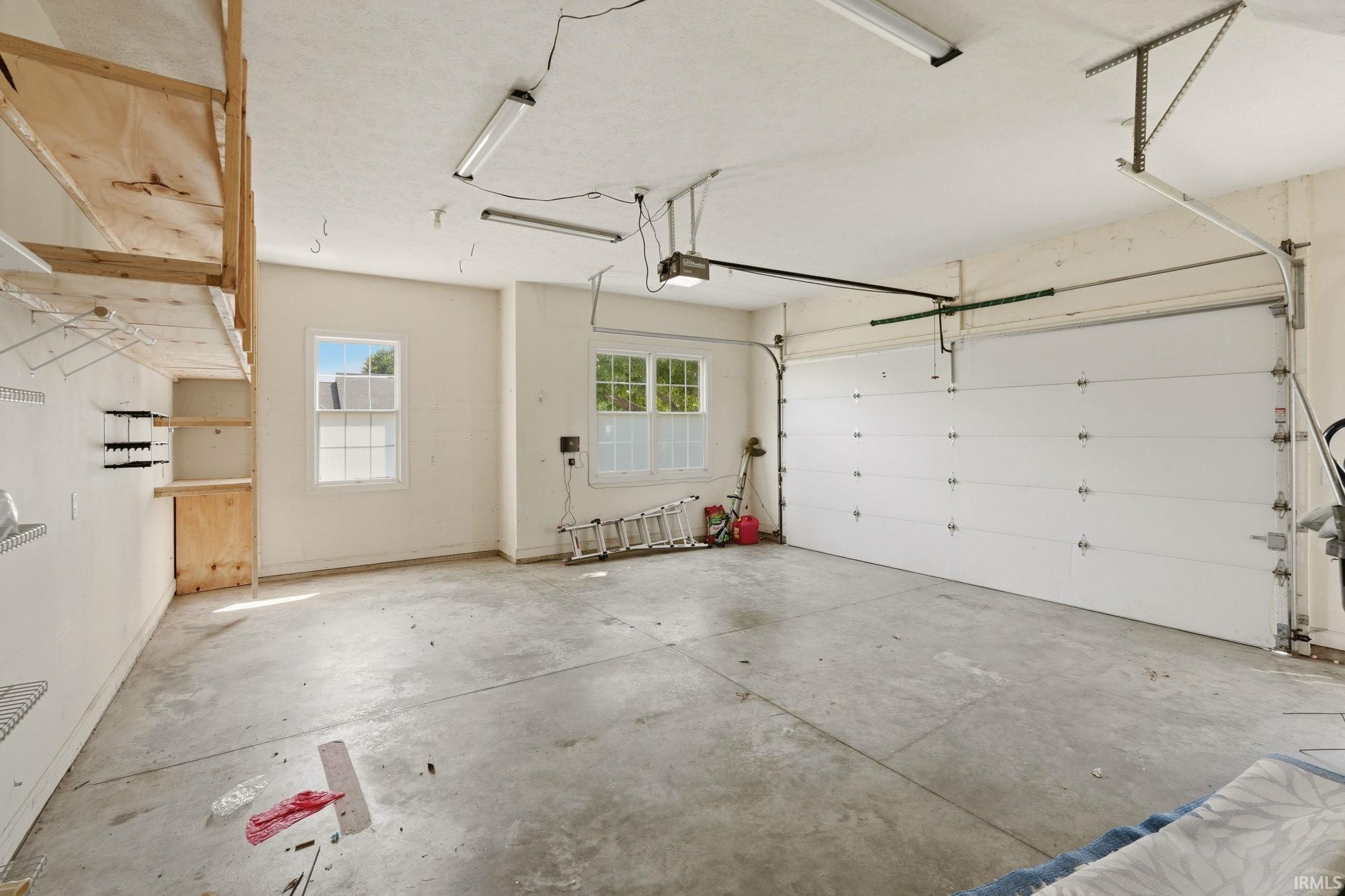 Spacious garage featuring a concrete floor, two windows providing natural light, a white paneled garage door, and built-in shelving with overhead storage