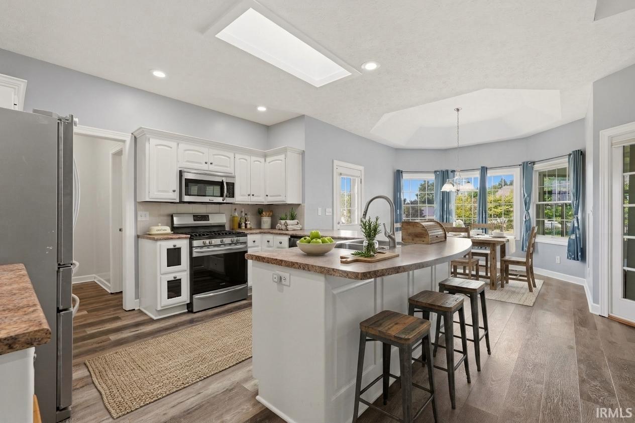 White cabinetry, stainless steel appliances, wood-finish flooring, kitchen island with seating, and a bay window dining area