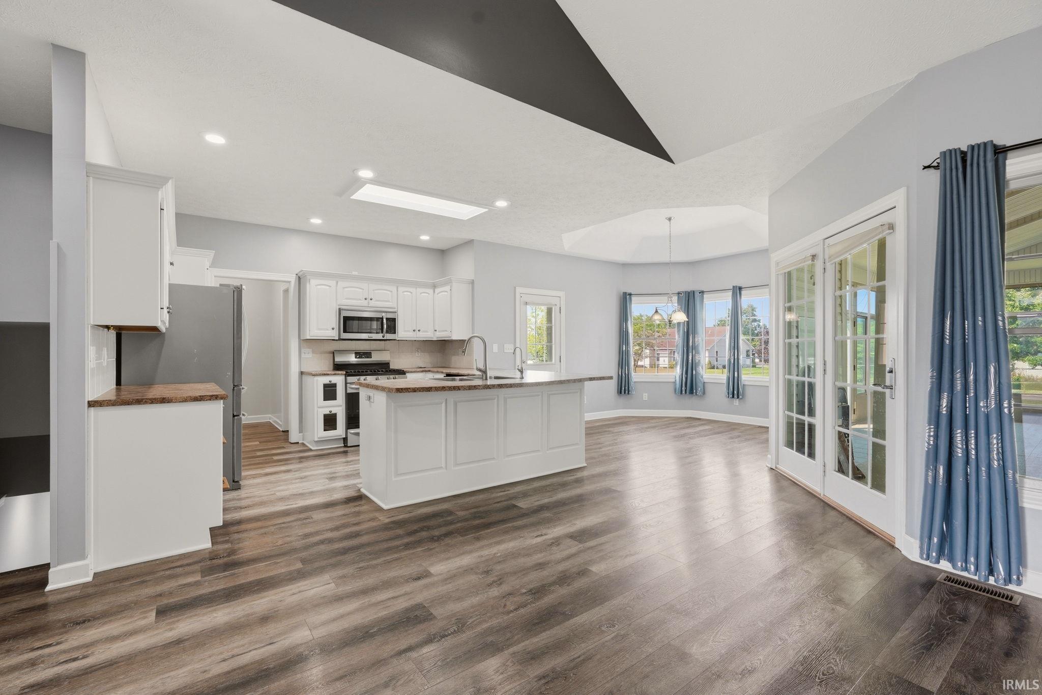 Open-concept kitchen and living area featuring wood-finish flooring, white cabinetry, a kitchen island with sink, stainless steel appliances, and a skylight