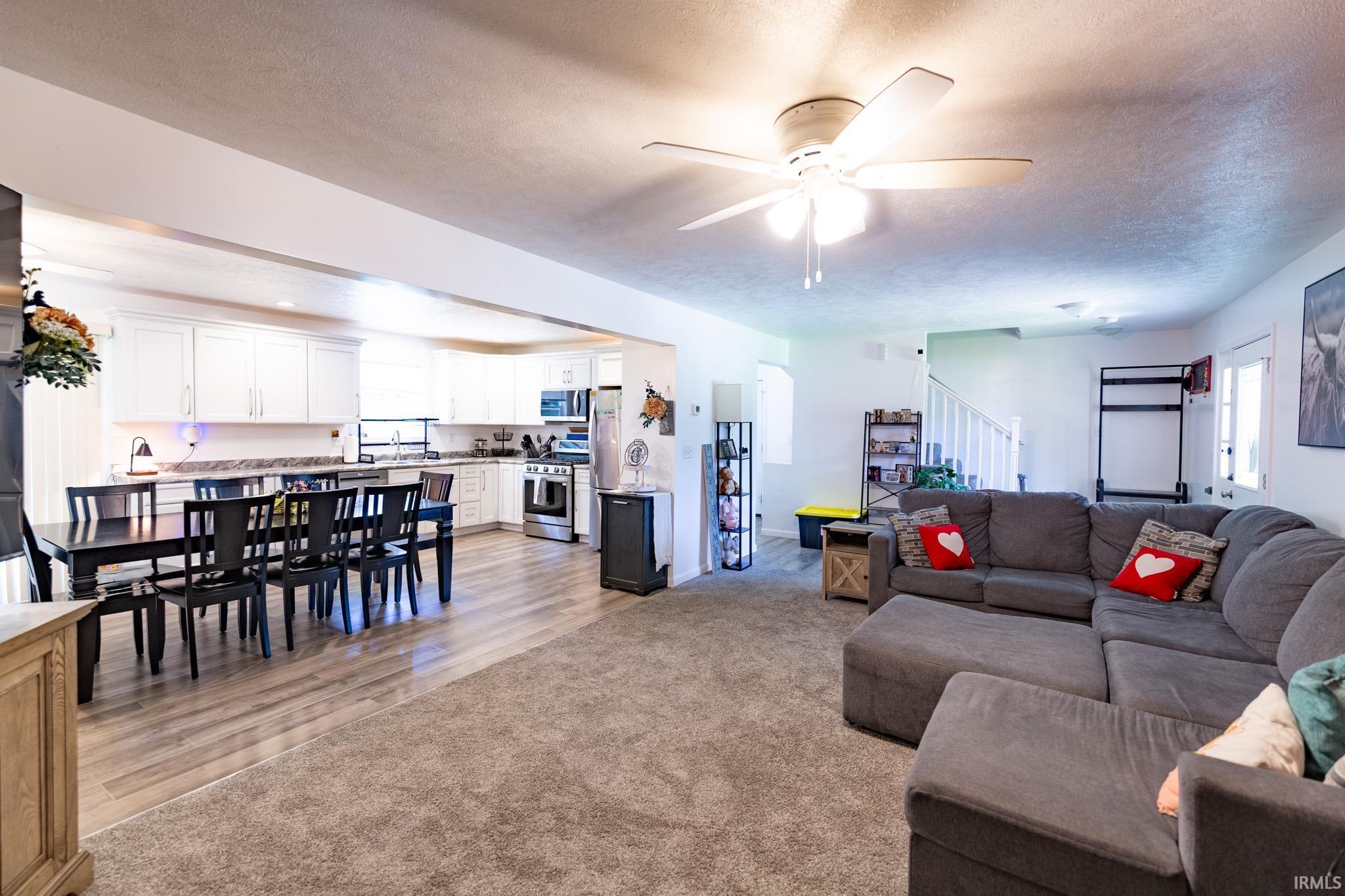 Open-concept living space featuring a spacious kitchen with white cabinetry and stainless steel appliances, a dining area with wood-finish flooring, and a carpeted living room