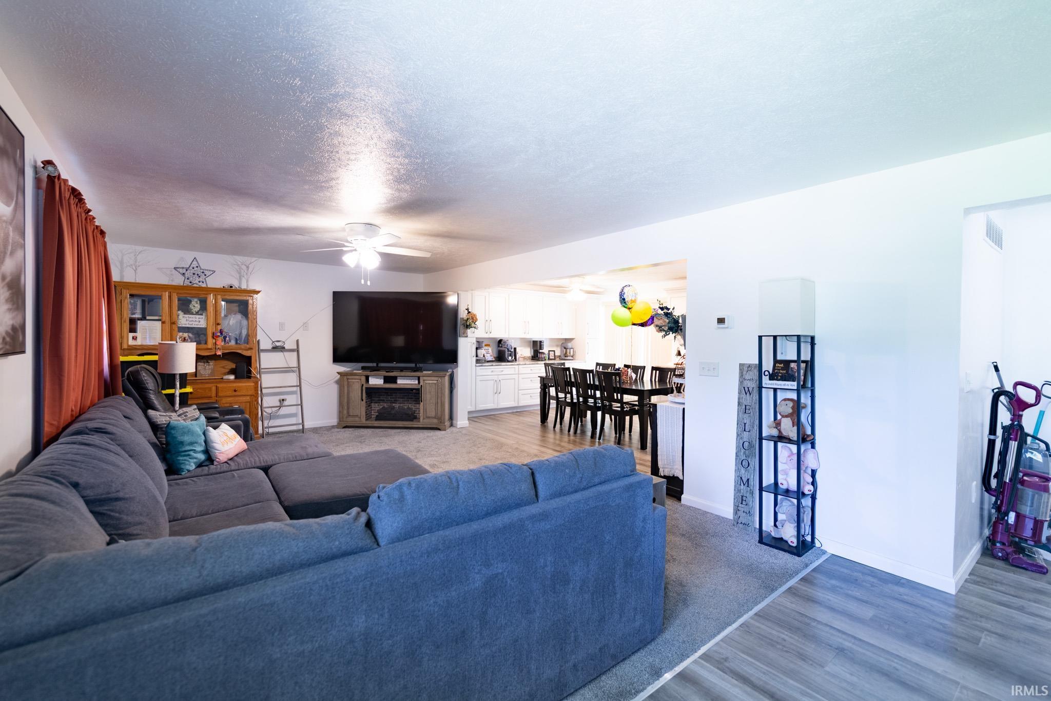 Carpeted living area with a ceiling fan, entertainment console, and wood-finish flooring extending into the kitchen and dining areas