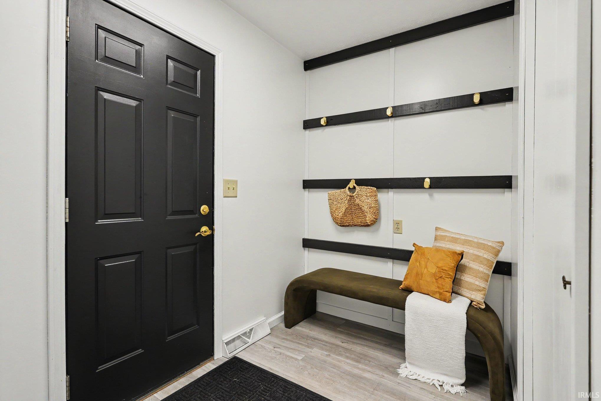 Entryway featuring a paneled black front door with brass hardware, light wood-finish flooring, and a white wall with black horizontal accents and brass hooks