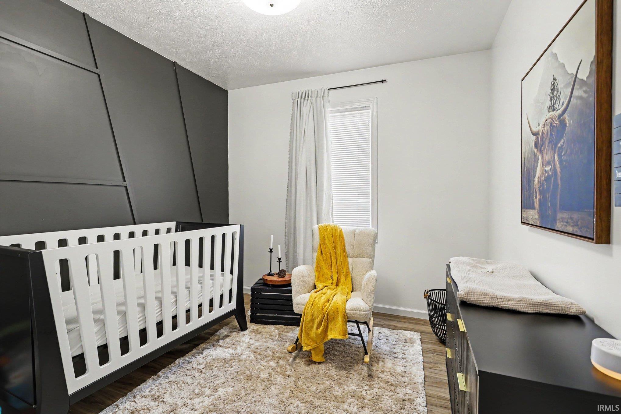 Bedroom featuring a geometric accent wall, wood-finish flooring, a flush-mount ceiling light, and a window with blinds and drapes