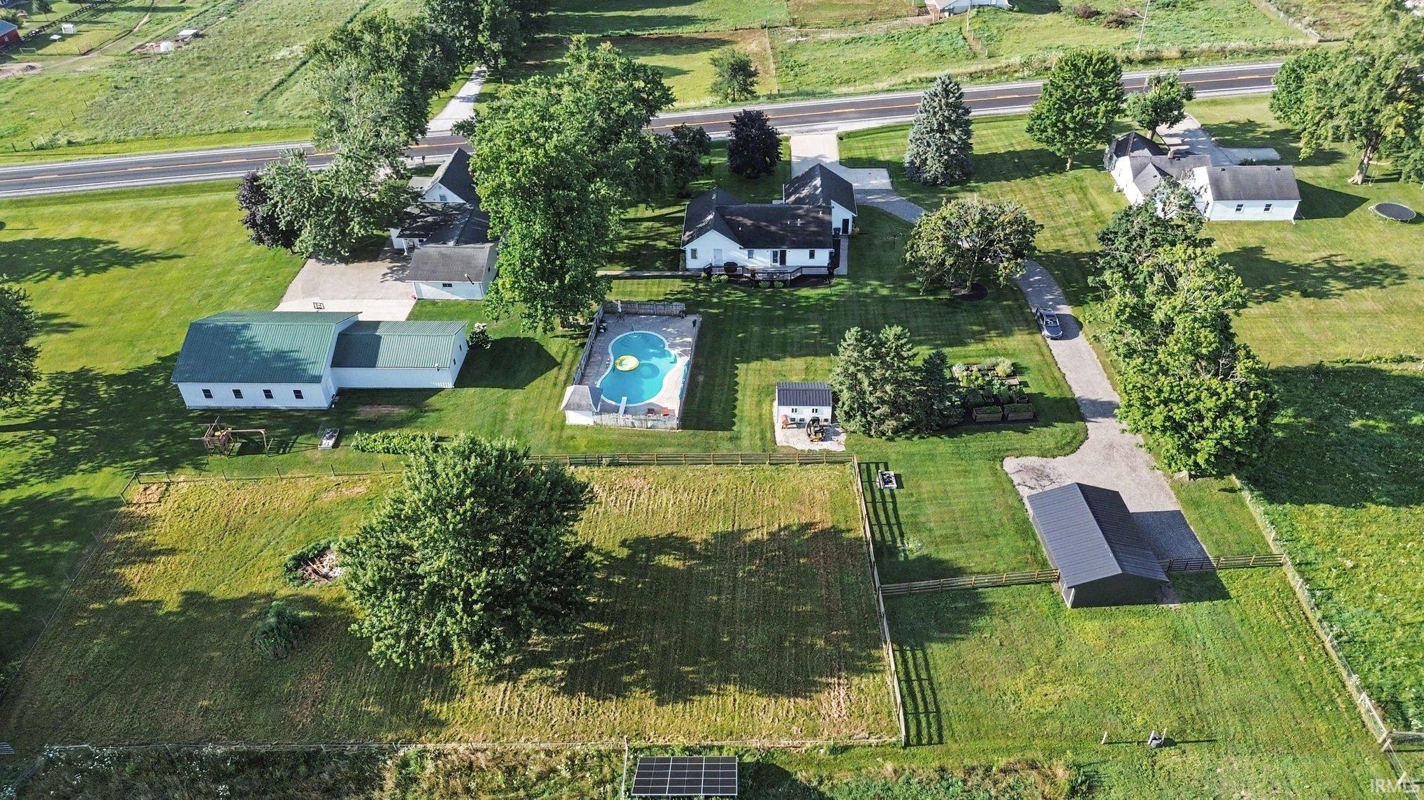Expansive property featuring multiple structures, a built-in swimming pool, a substantial detached outbuilding with a dark roof, and mature perimeter trees