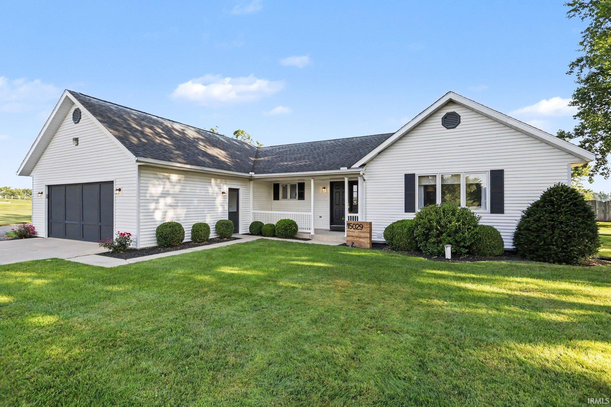 White siding exterior with a dark-paneled garage door, black window shutters, a covered porch entry, and a manicured front lawn