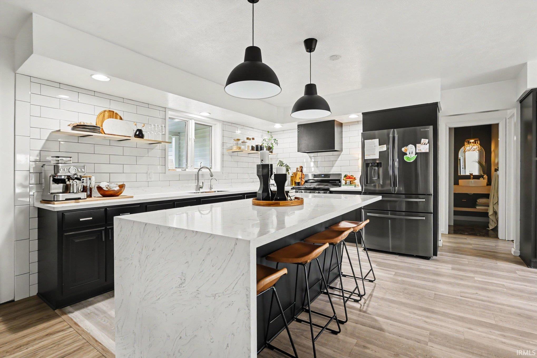 Modern kitchen featuring a large waterfall island, white subway tile backsplash, dark cabinetry, wood-finish flooring, and pendant lighting