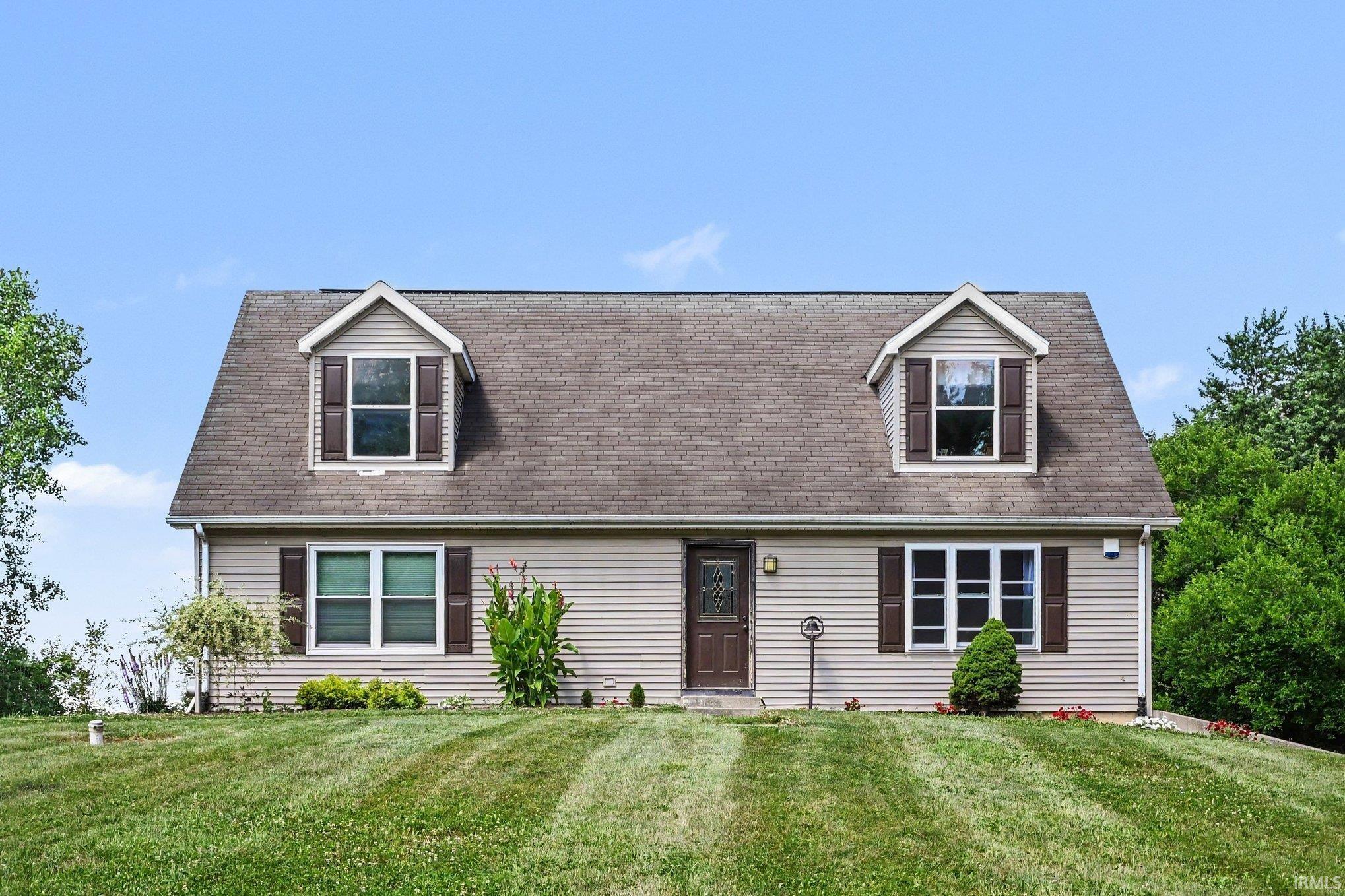 Dormered Cape Cod style residence featuring beige siding, dark brown entry door, dark brown window shutters, and a sloped front yard