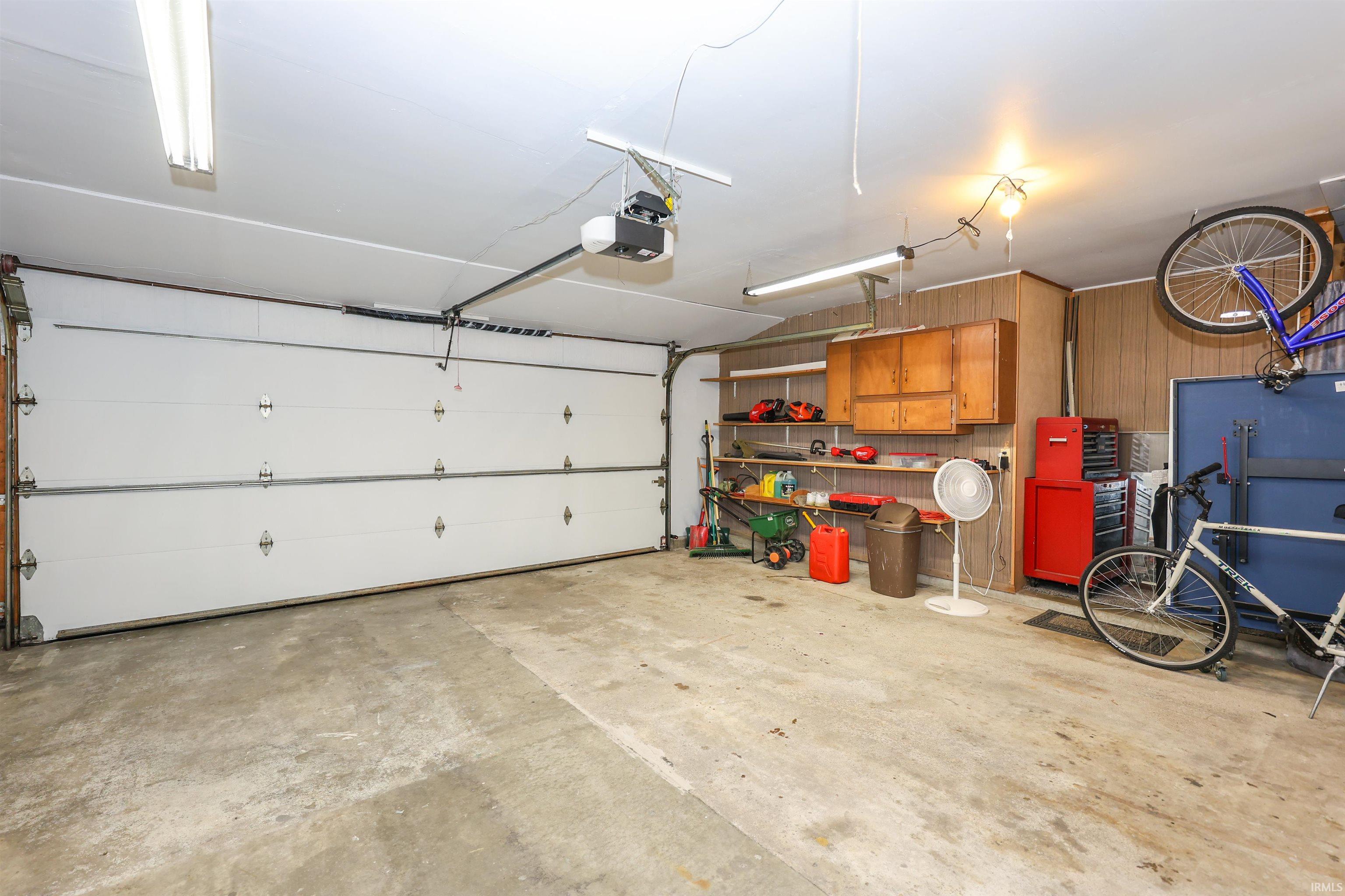Two-car garage with concrete flooring, overhead fluorescent lighting, wood paneling, and built-in shelving