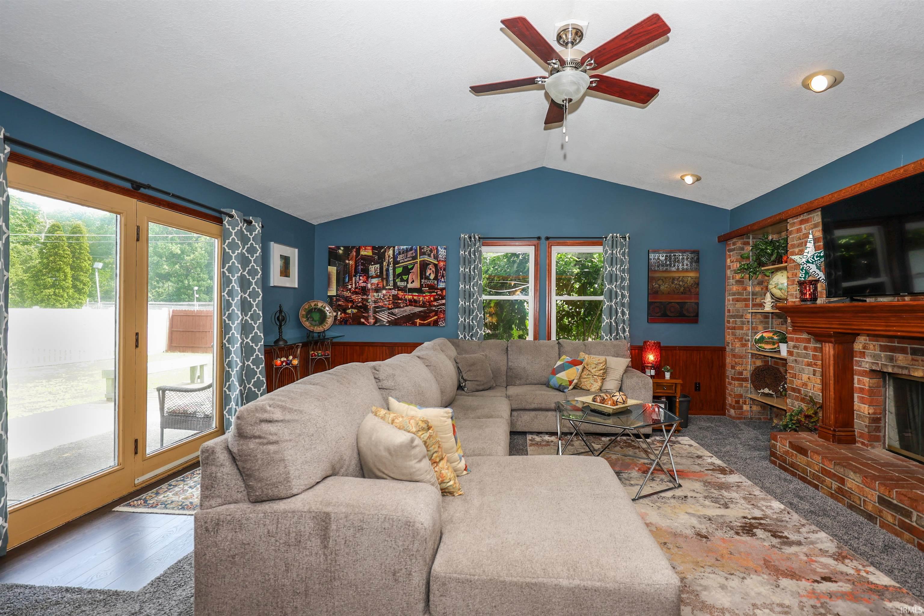 Spacious living area featuring a vaulted ceiling with a ceiling fan, brick fireplace with a wood mantel, and wood-finish paneling