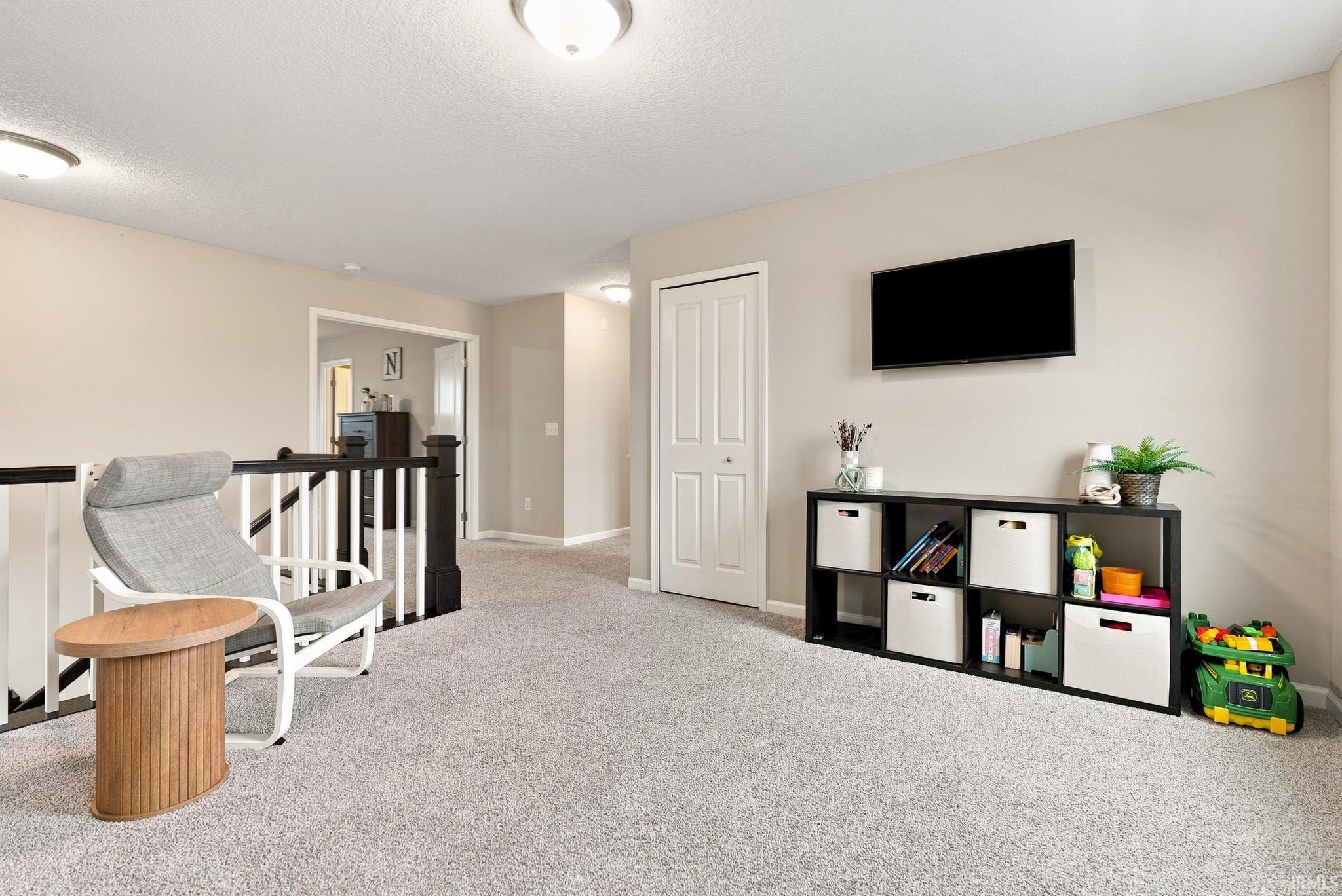 Carpeted loft area featuring a wall-mounted display, neutral wall tones, white baseboards, and a bi-fold door