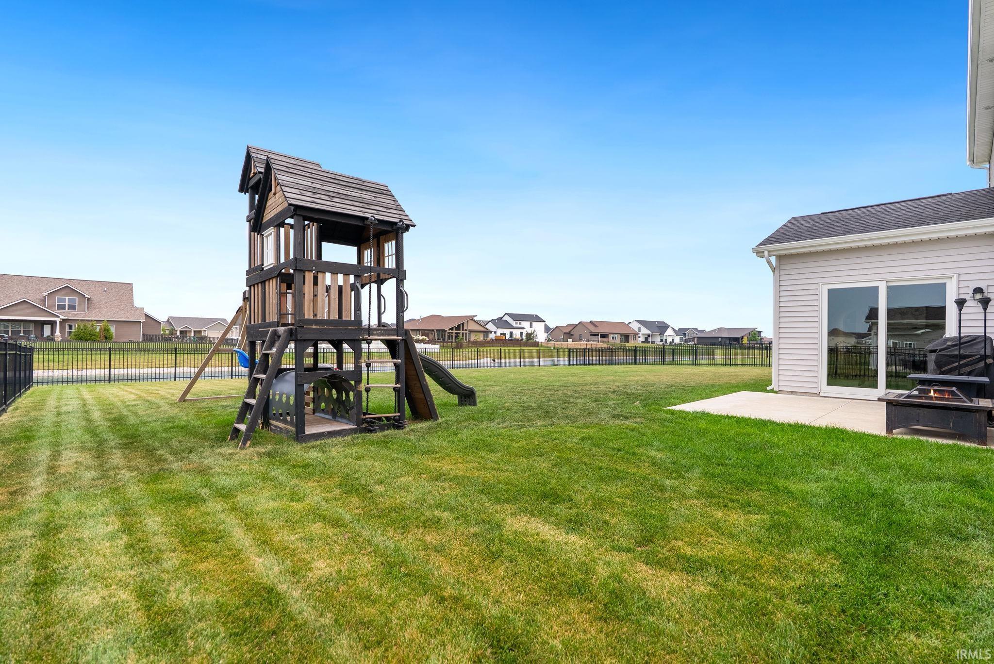 Expansive rear lawn featuring a wooden playset with slide and swings, a concrete patio, and a black metal fence