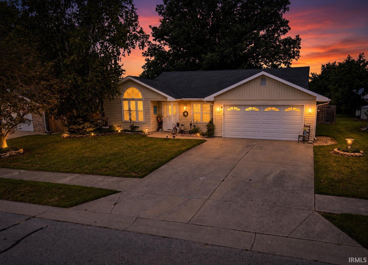Siding exterior featuring a prominent arched window, attached garage with decorative paneled door, concrete driveway, and front lawn with mature trees and integrated lighting