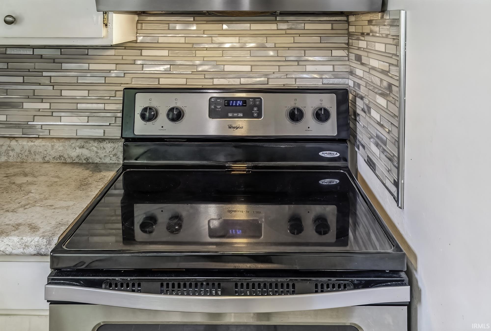 Stainless steel range with a black ceramic cooktop, accompanied by a glass tile backsplash and light-toned, speckled countertops