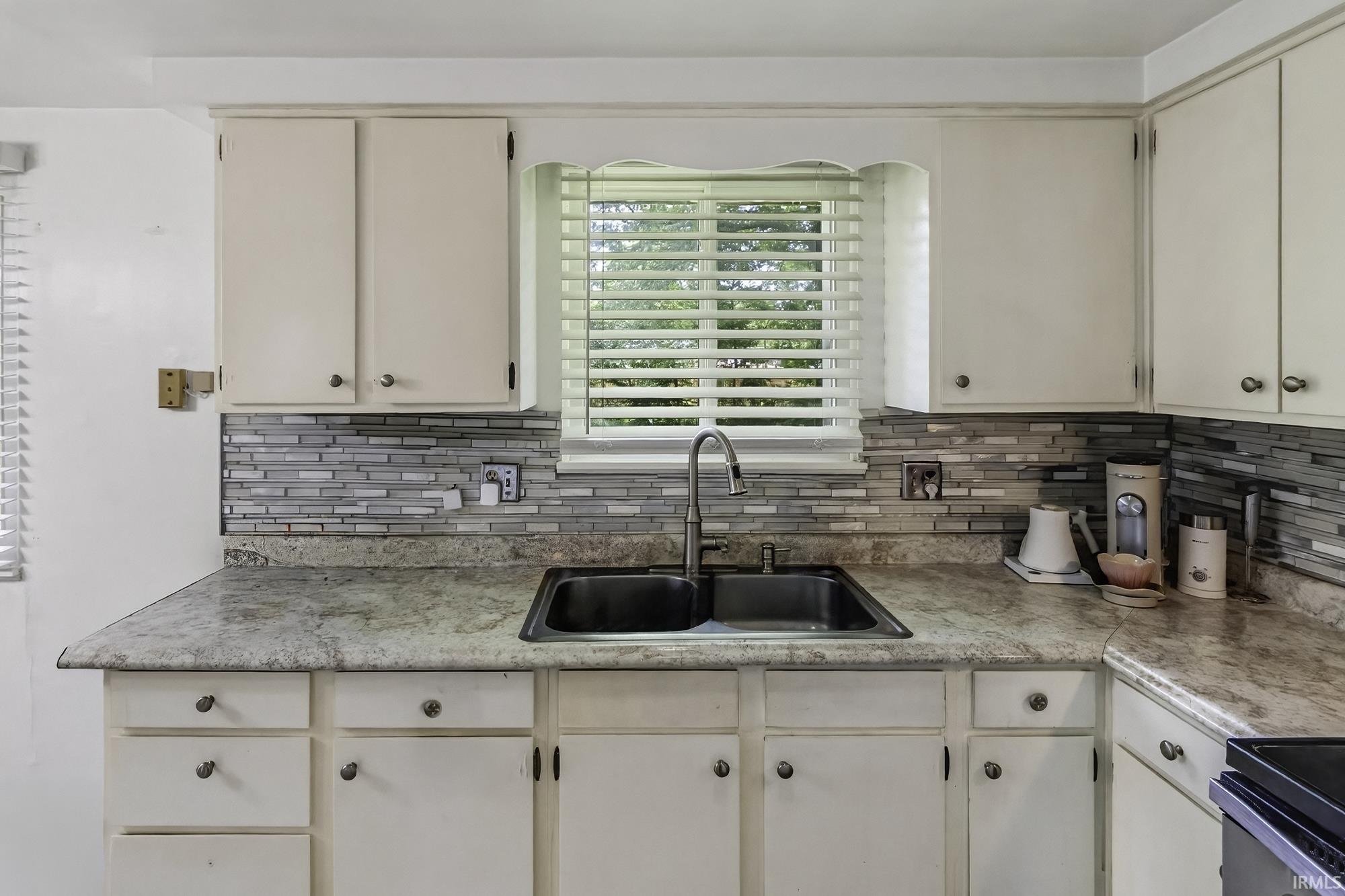 Kitchen featuring a double basin sink with a gooseneck faucet, light-toned countertops, white cabinetry, and a mosaic tile backsplash