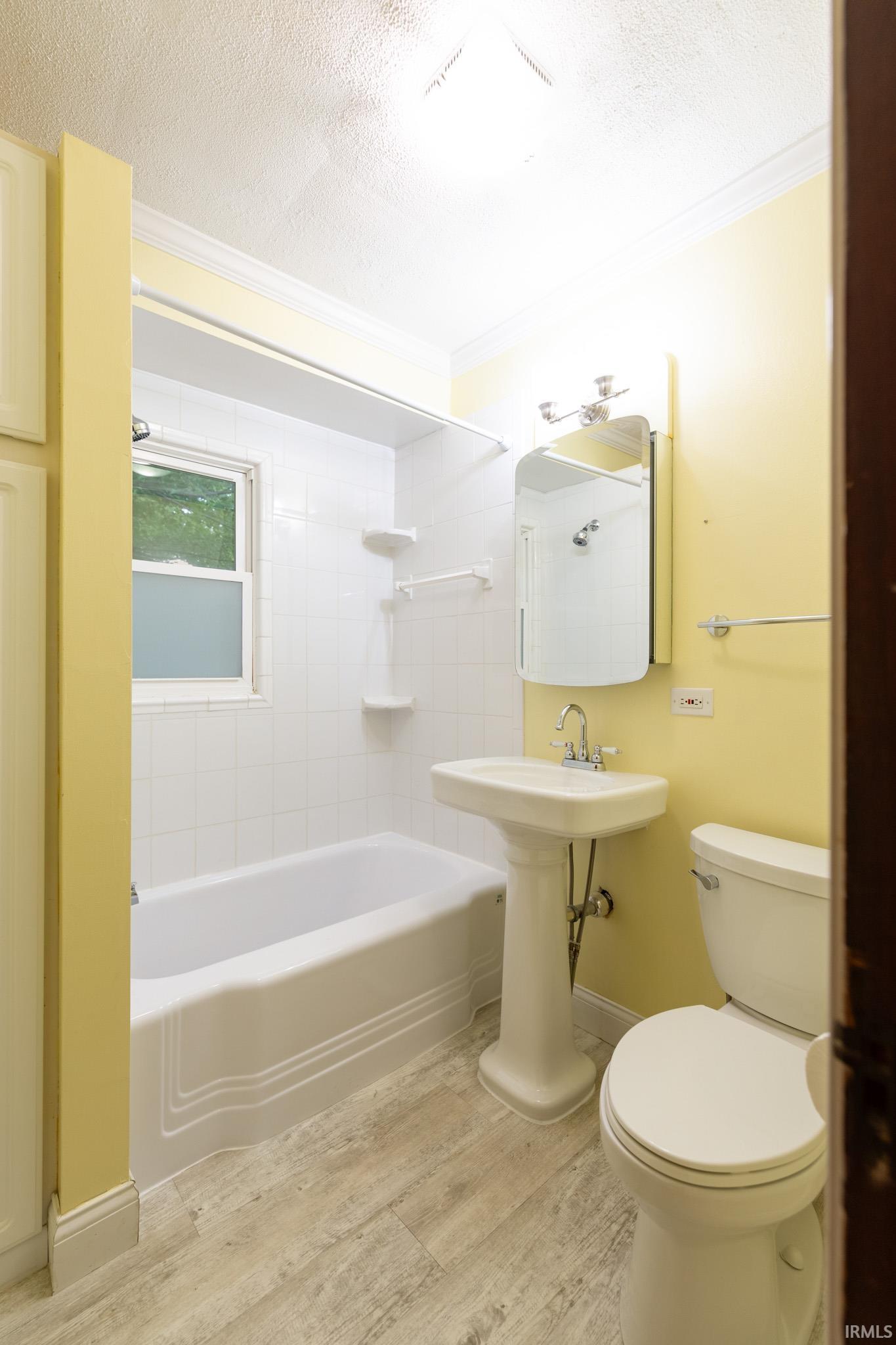 Bathroom featuring white pedestal sink, white tile shower surround with integrated tub, wood-finish flooring, and a window with frosted glass