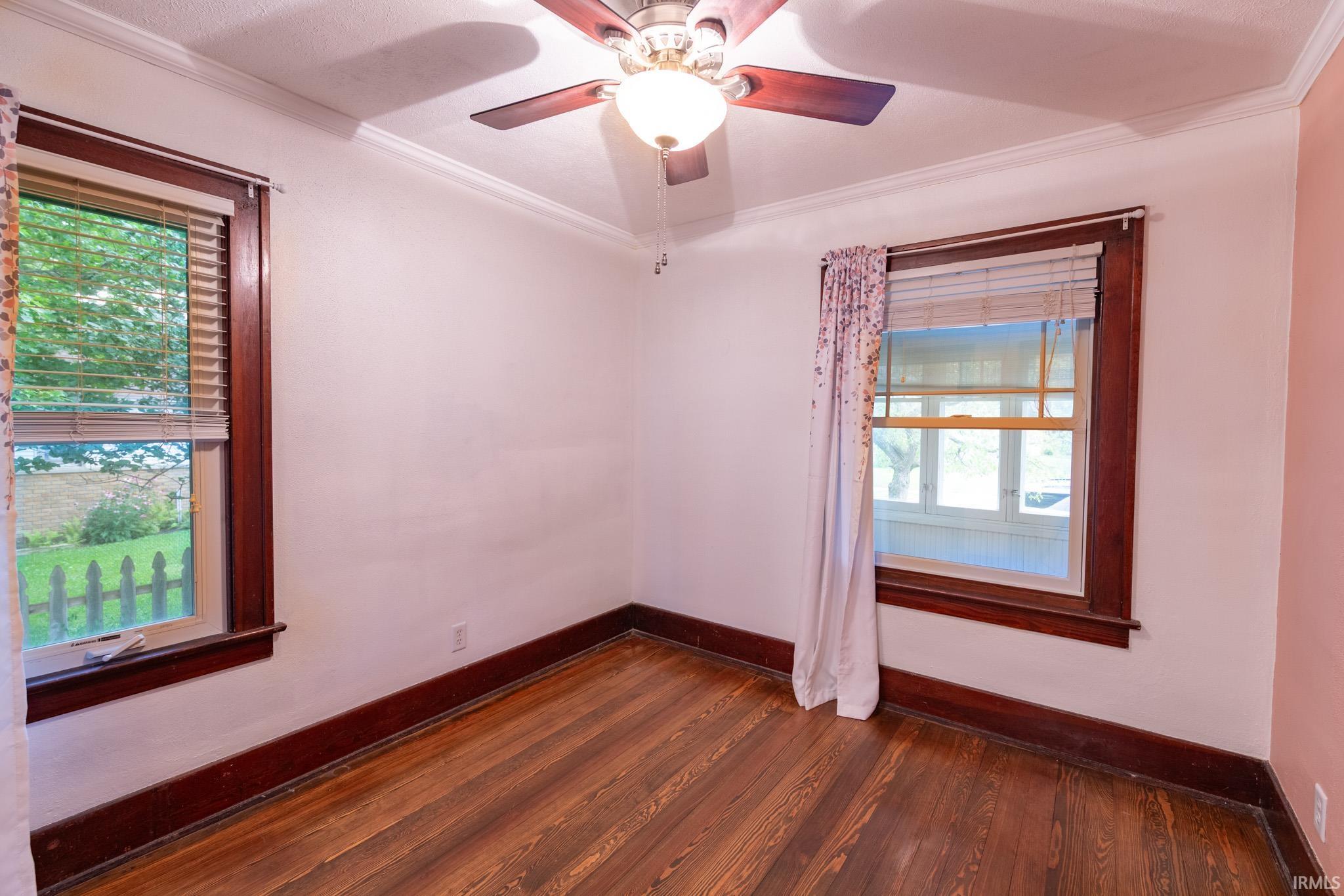 Room featuring wood-finish flooring, two windows with dark wood trim, crown molding, and a ceiling fan with integrated lighting