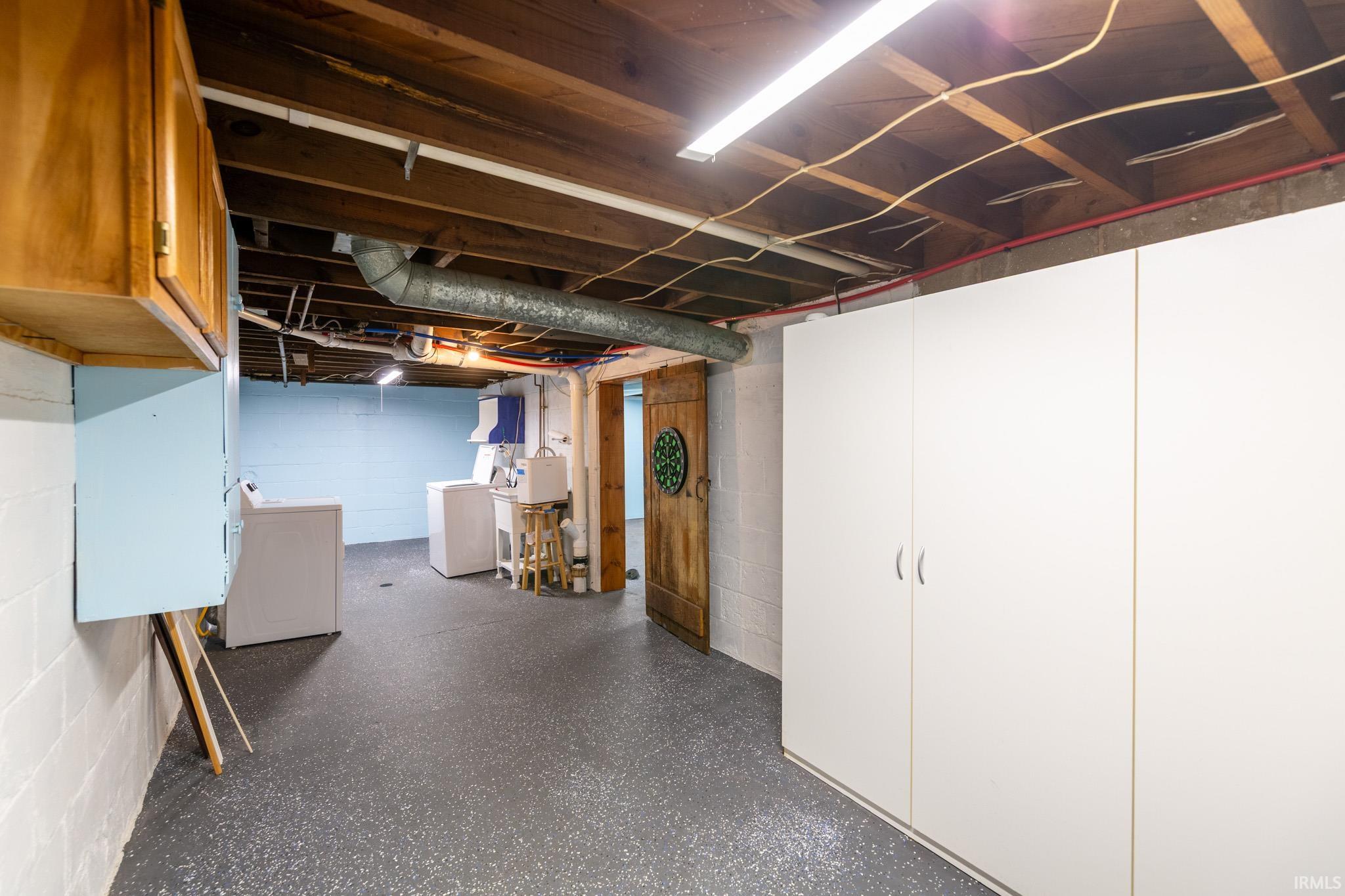 Basement utility area featuring exposed joists, bright fluorescent lighting, extensive built-in white cabinetry, a utility sink, and a wooden door