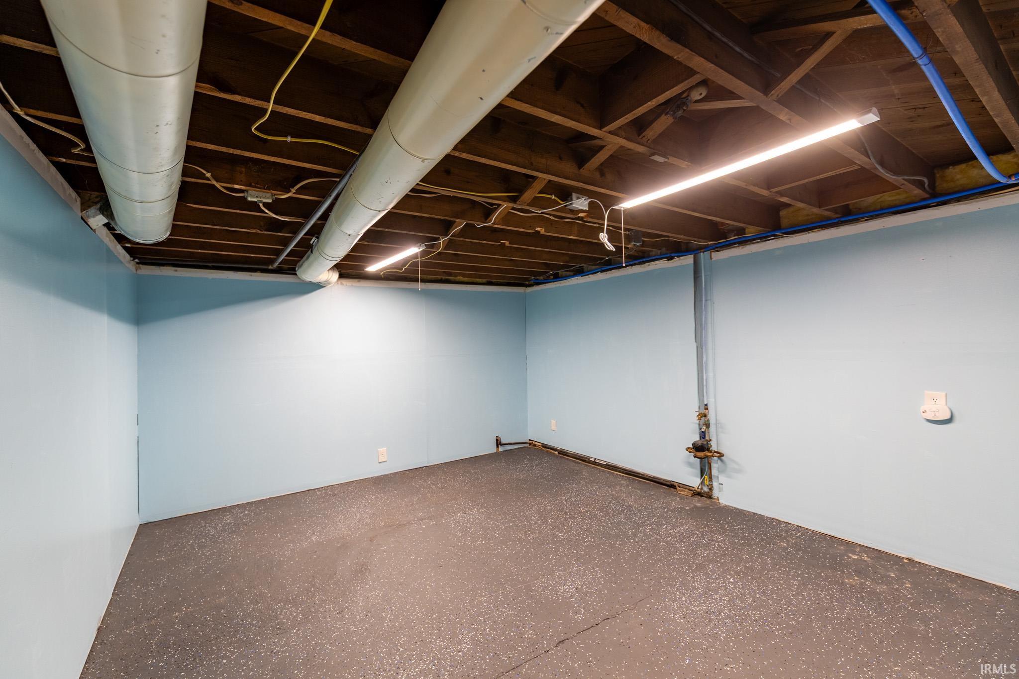 Finished basement room with light blue painted walls, exposed ceiling joists, durable gray speckled flooring, and linear fluorescent lighting fixtures