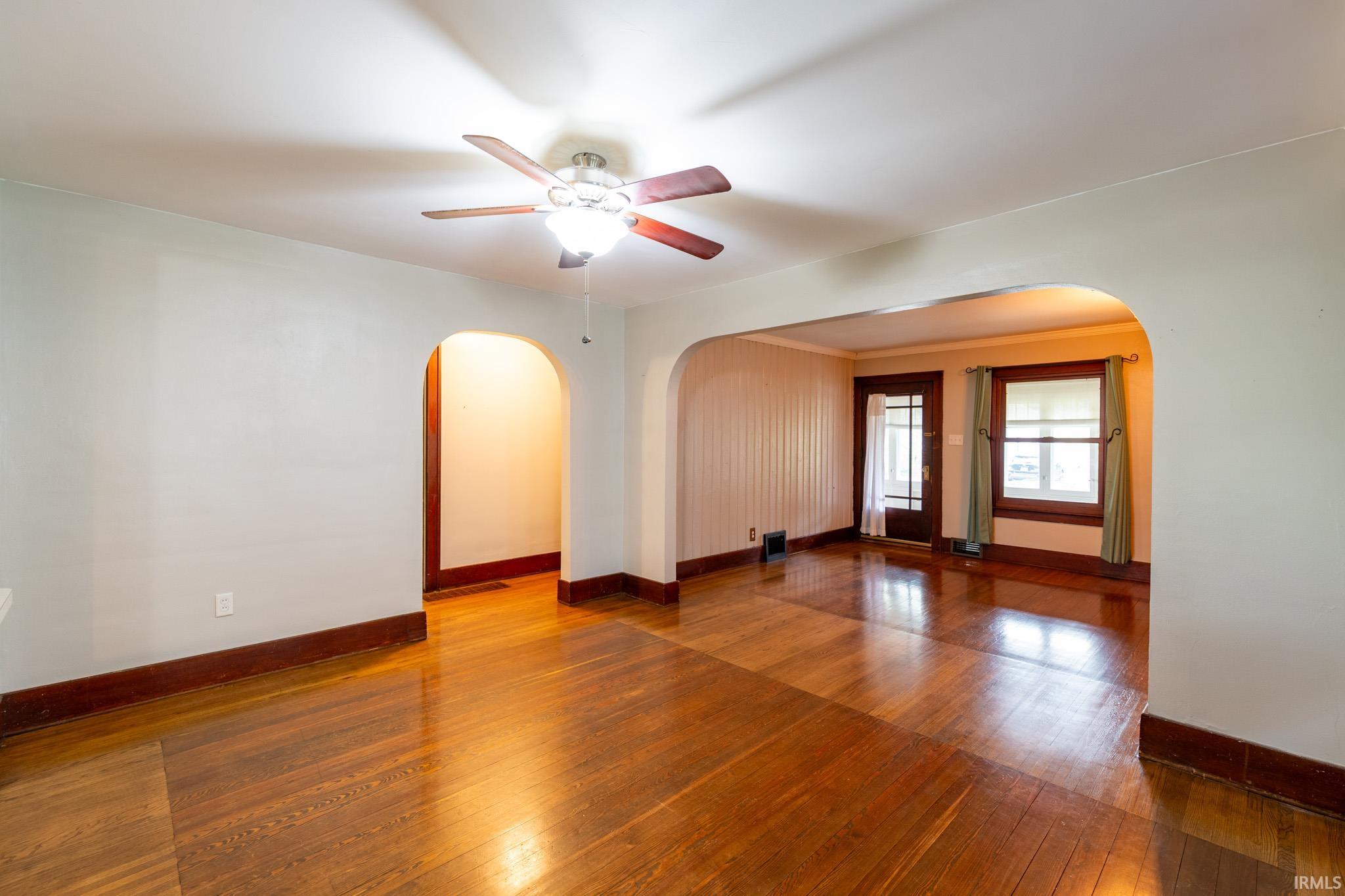 Hardwood flooring extends throughout this interior space featuring arched doorways, a ceiling fan with integrated lighting, and multiple windows with dark wood trim