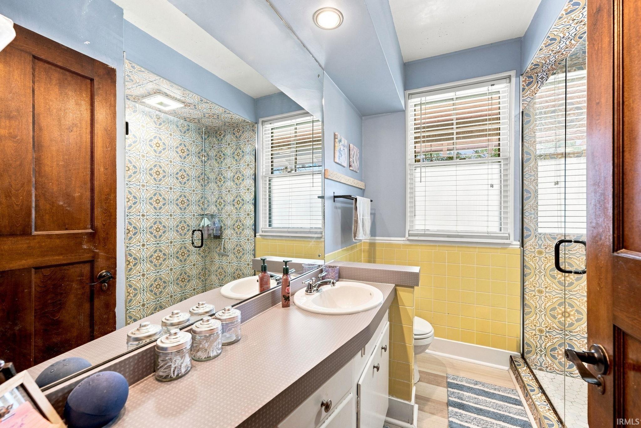 Bathroom featuring a dual-sink vanity with white basin sinks, patterned tile shower enclosure with glass door, two windows with blinds, and yellow wall tiling
