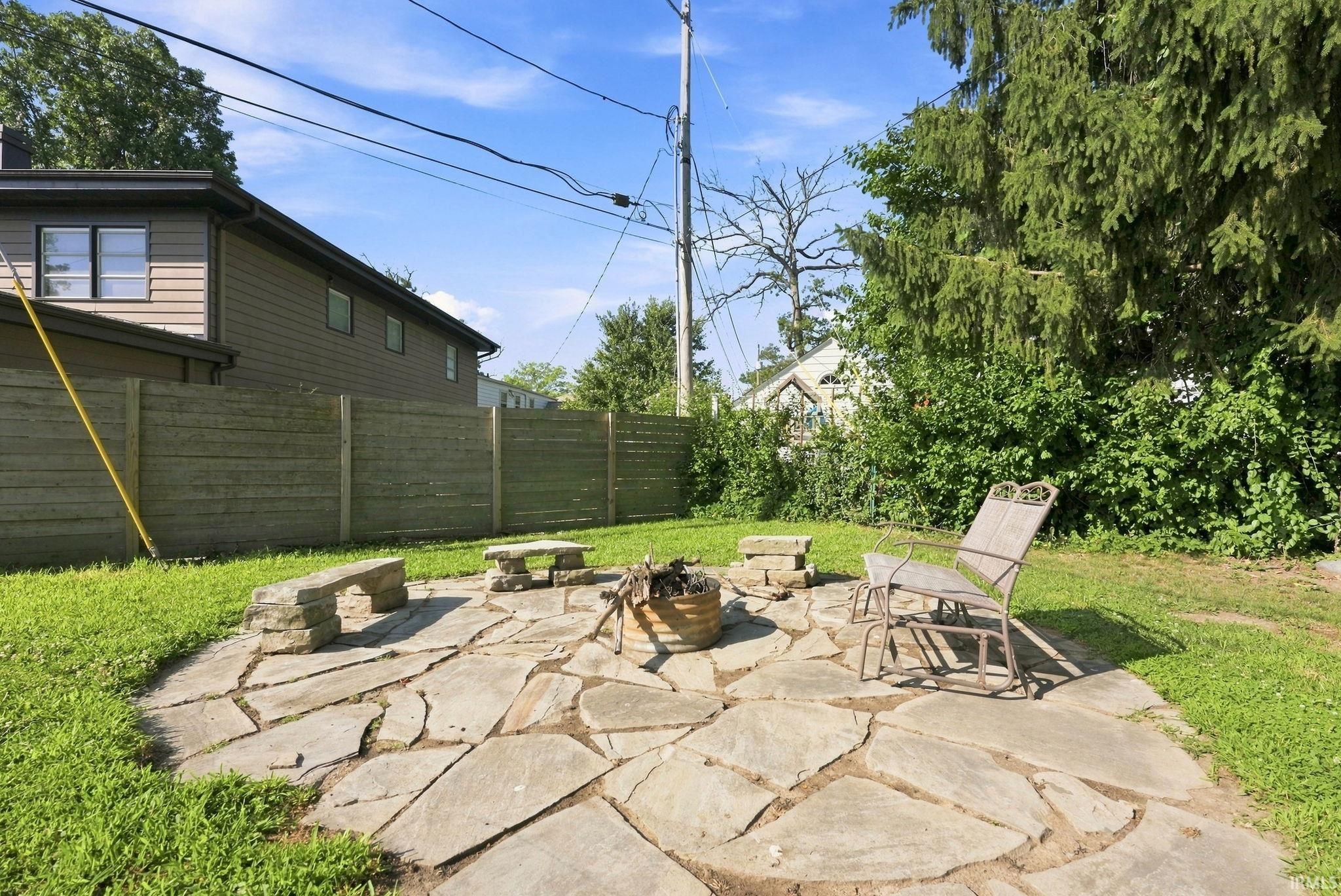 Stone patio featuring a central fire pit, surrounded by a manicured lawn and privacy fencing