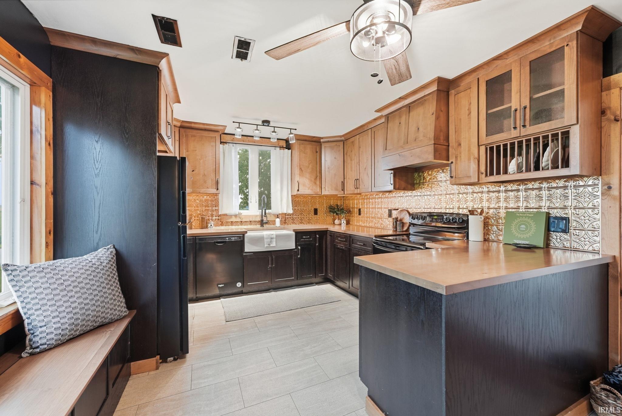 Kitchen featuring wood-finish cabinetry, black base cabinetry, a white apron-front sink, an ornate backsplash, and a ceiling fan