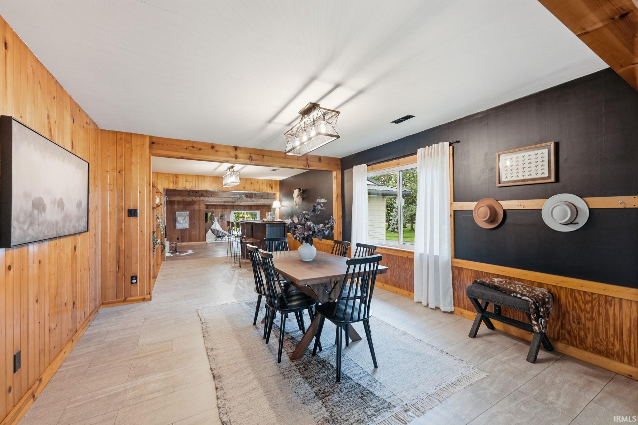 Wood paneling throughout, exposed wood beam, rectangular window with white trim, multi-light linear chandelier, and light-toned flooring